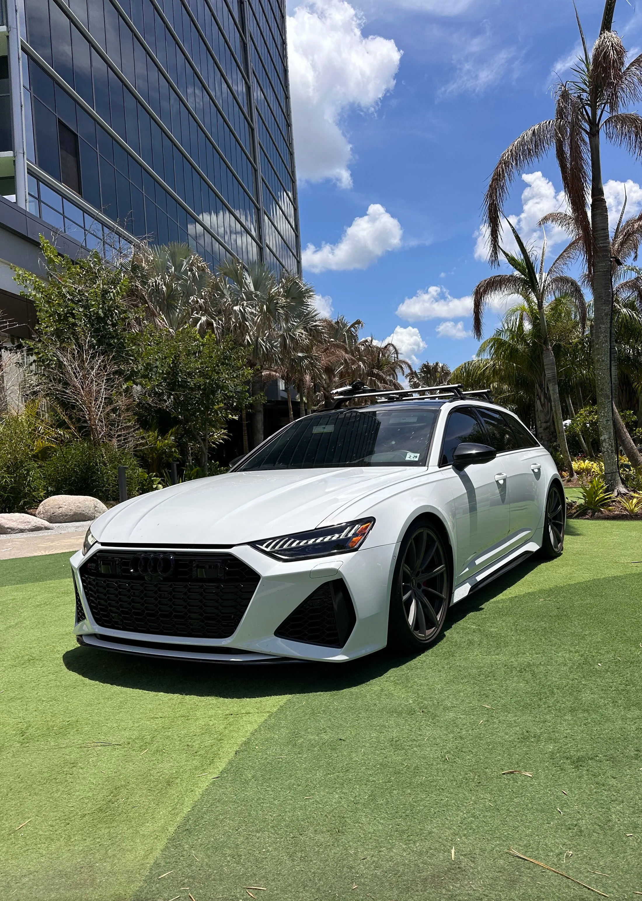A white sports car parked on a green synthetic grass surface outdoors, with a modern glass building, palm trees, and blue sky with clouds in the background.