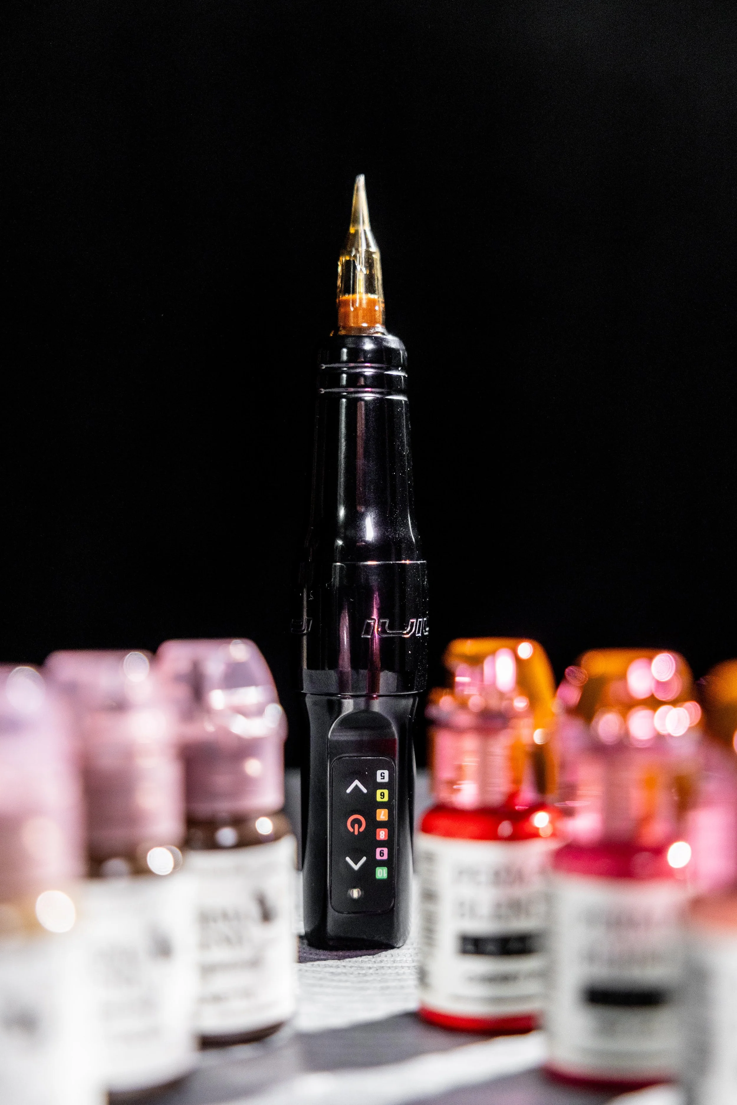Close-up of a tattoo machine with ink bottles in the foreground against a black background.