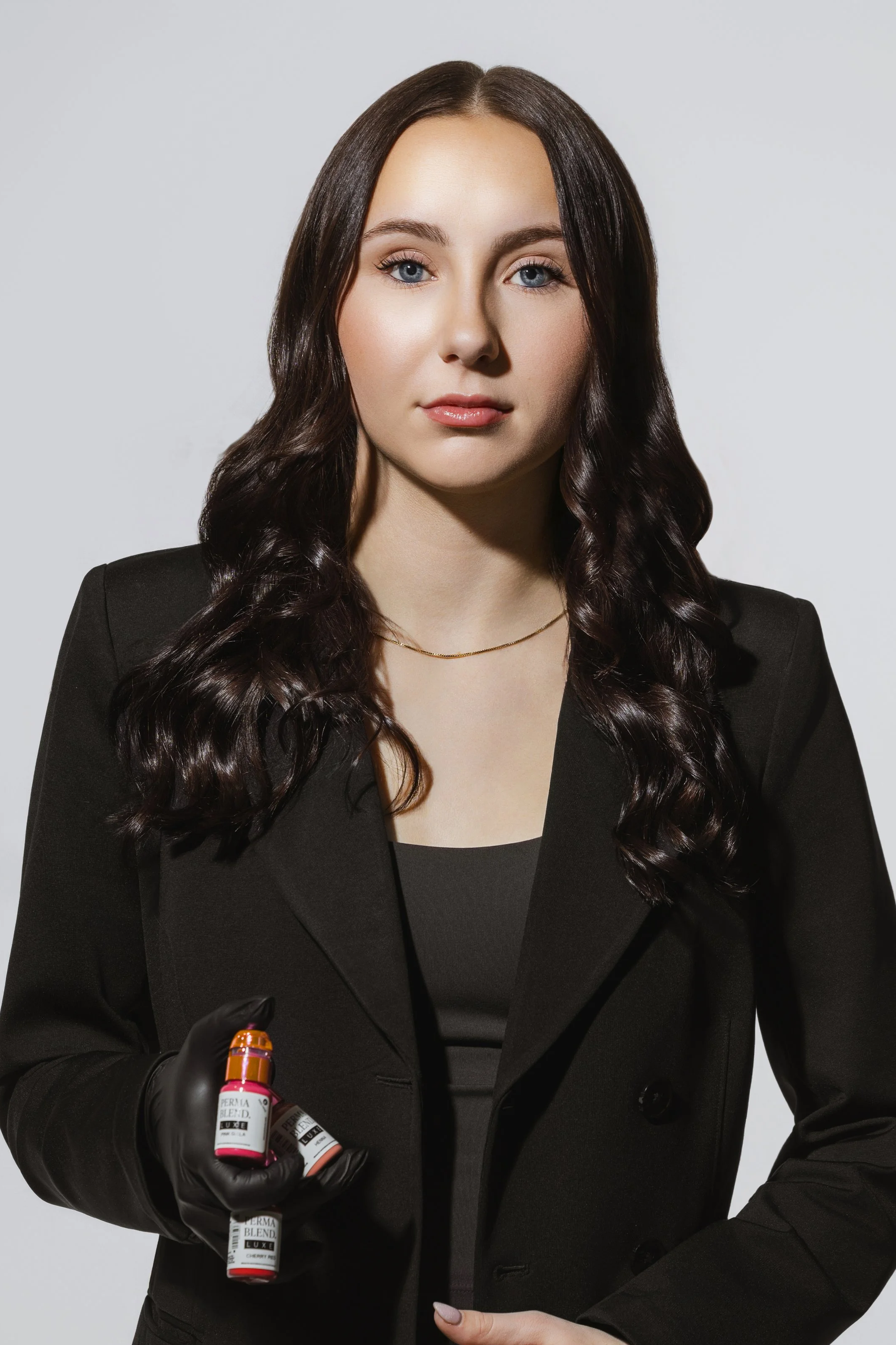 A woman with dark wavy hair in a black suit holding bottles of Perry Bleach hair dye, wearing black gloves, against a plain light gray background.