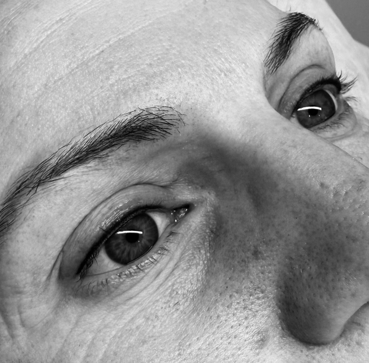 Close-up black and white photograph of a person's face showing only the eyes and part of the nose, with detailed skin texture and eyebrows.