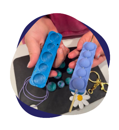 Person holding two slap bracelets with finger slime or putty in blue and purple, with a white flower-shaped item and gold keychain in the background.