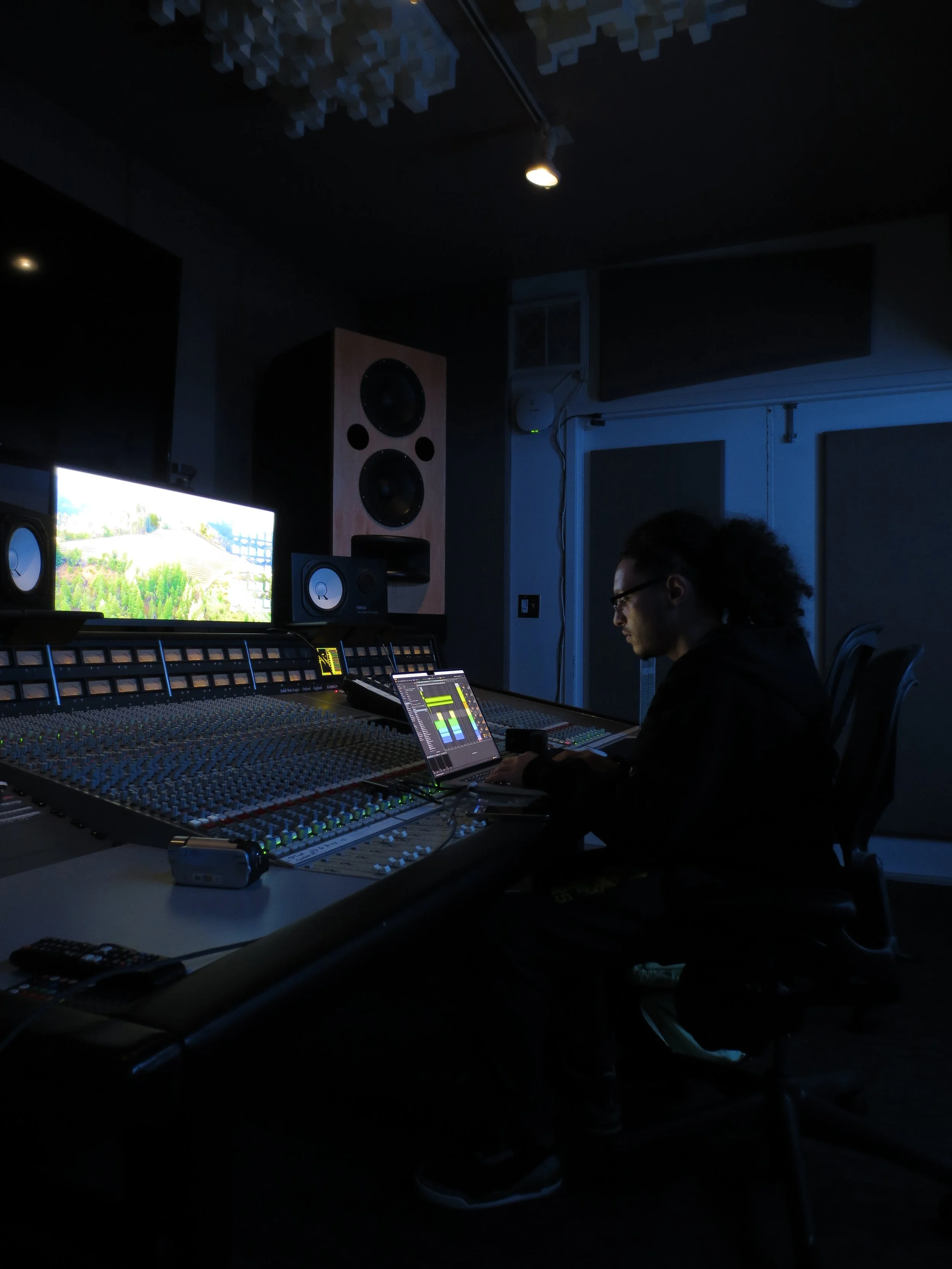 A person working in a recording studio at a mixing console with a computer monitor and speakers, dark lighting.