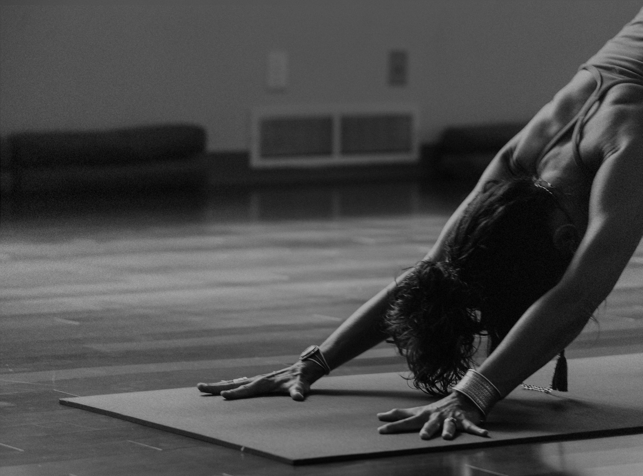 Black and white photo of a woman in a downward dog yoga pose on a mat in a room with wood floors, couches, and a heater in the background.