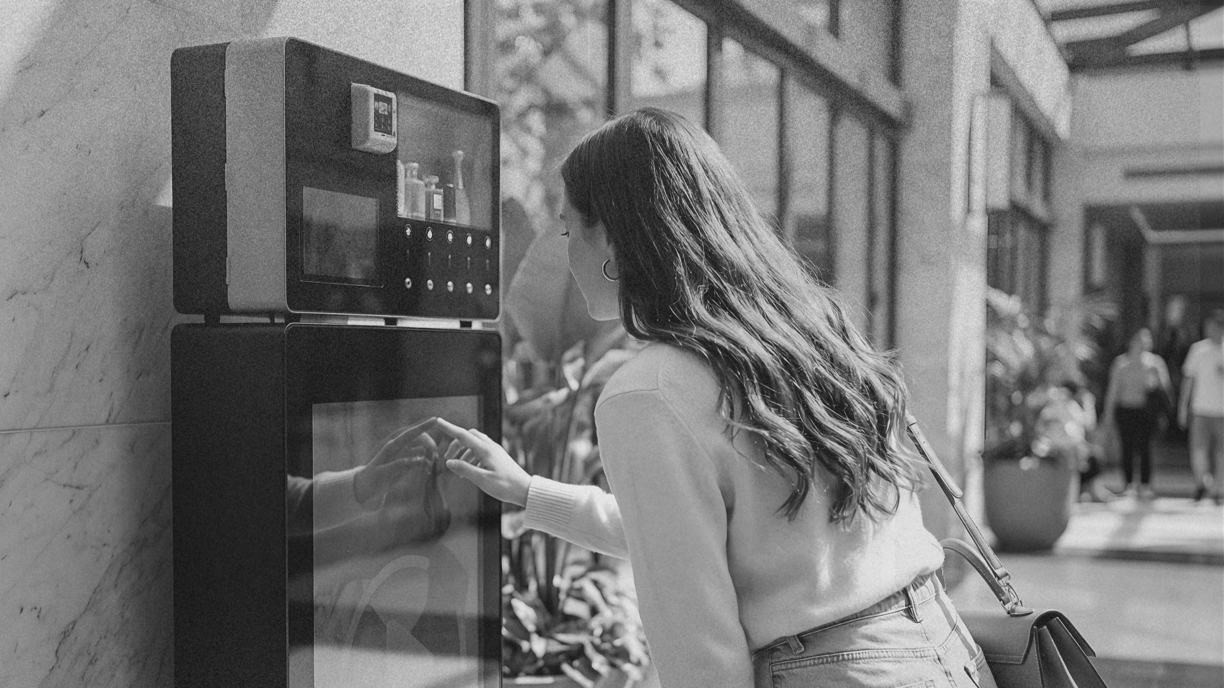 A young woman with long wavy hair, wearing a light-colored sweater and jeans, using a vending machine outdoors on a sunny day.