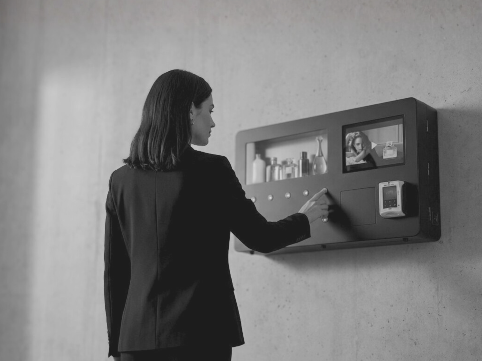 Woman in black blazer interacting with a perfume vending machine panel with a small display screen and various items inside a cabinet, against a plain wall. untilisation de l'écran tactile d'un Distributeur automatique de parfum.