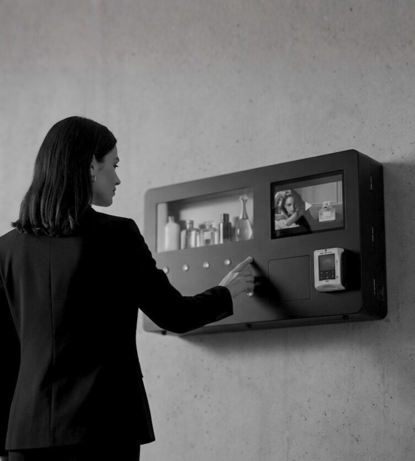 A woman in a black blazer using a touchscreen interface on a wall-mounted device, with a small screen showing her reflection, on a plain wall.