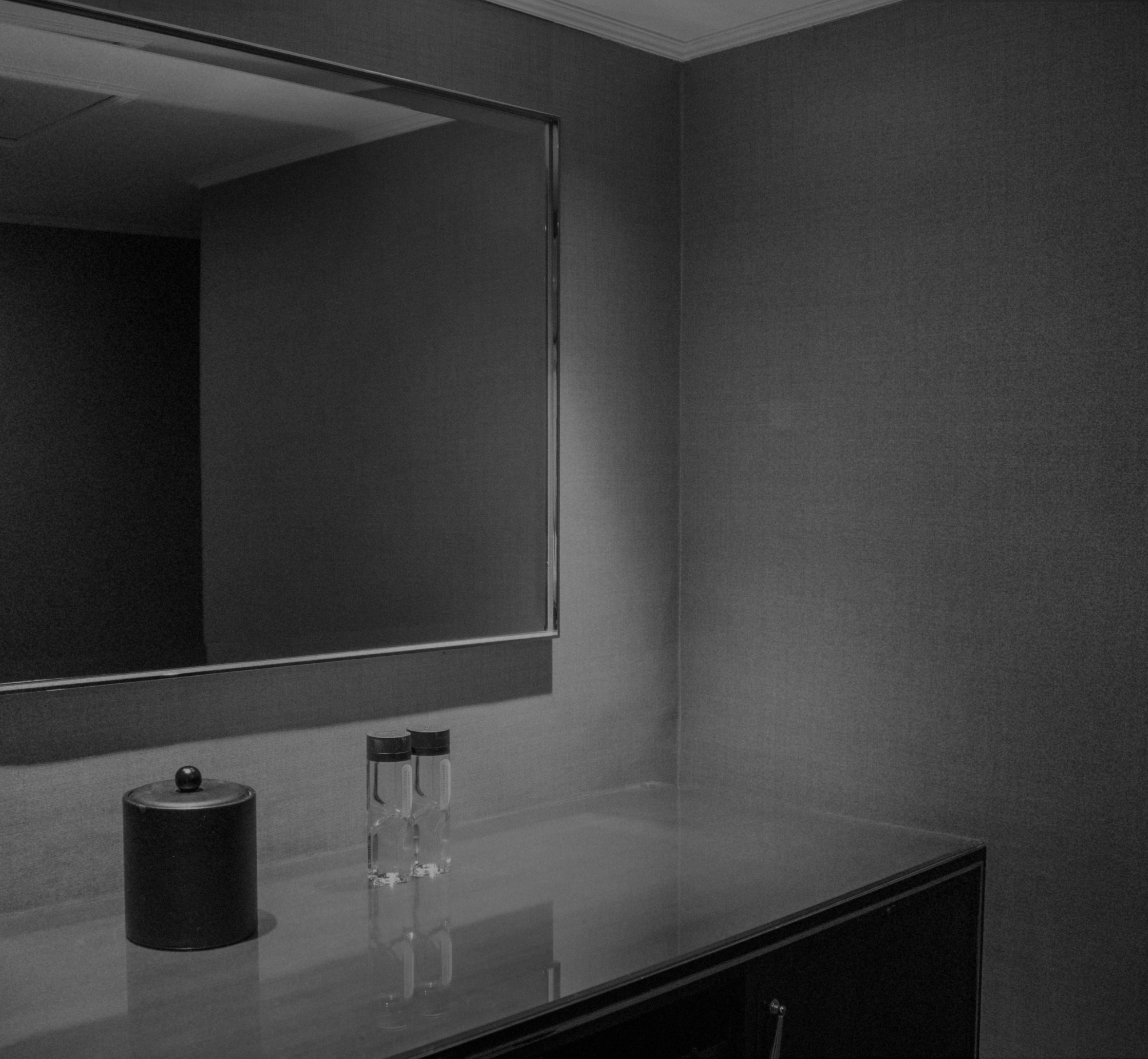 A black-and-white image of a bathroom vanity with a mirror, a cylindrical container with a lid, and two small bottles with black caps on a countertop.
