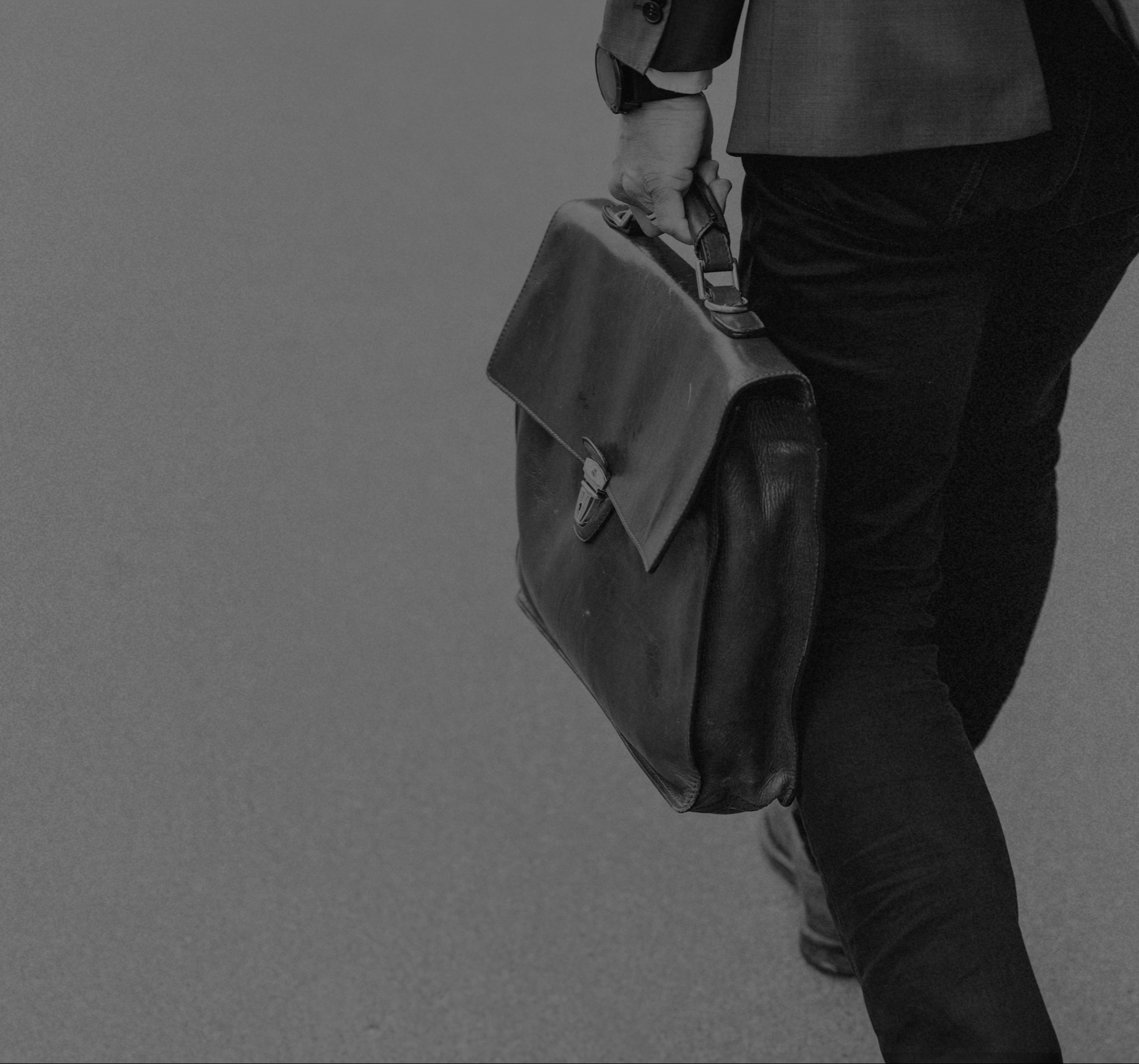 Businessman walking, carrying a leather briefcase in his right hand, dressed in a dark suit and black pants.