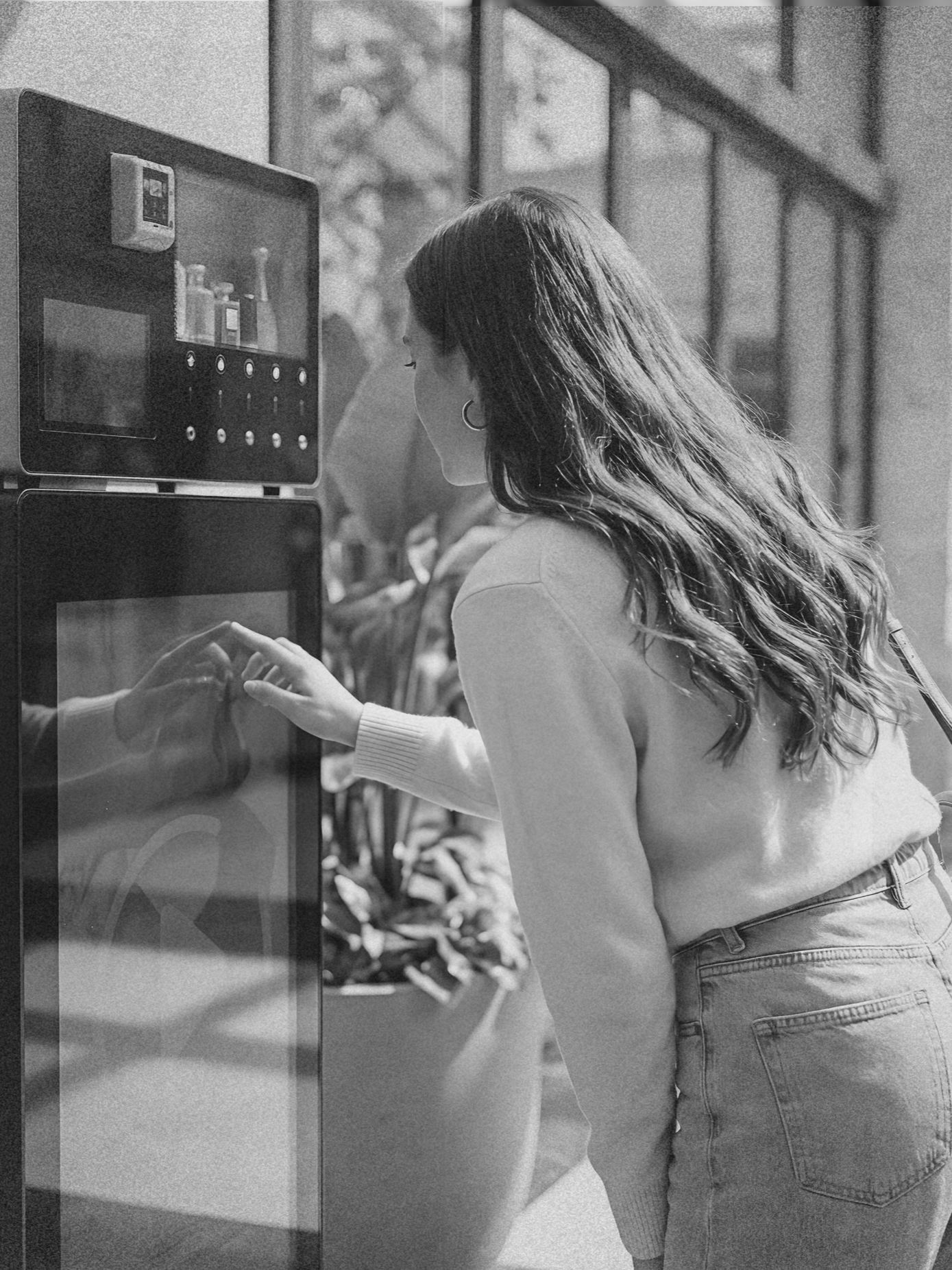 A woman with wavy hair wearing a light-colored sweater and jeans is selecting food at a vending machine.