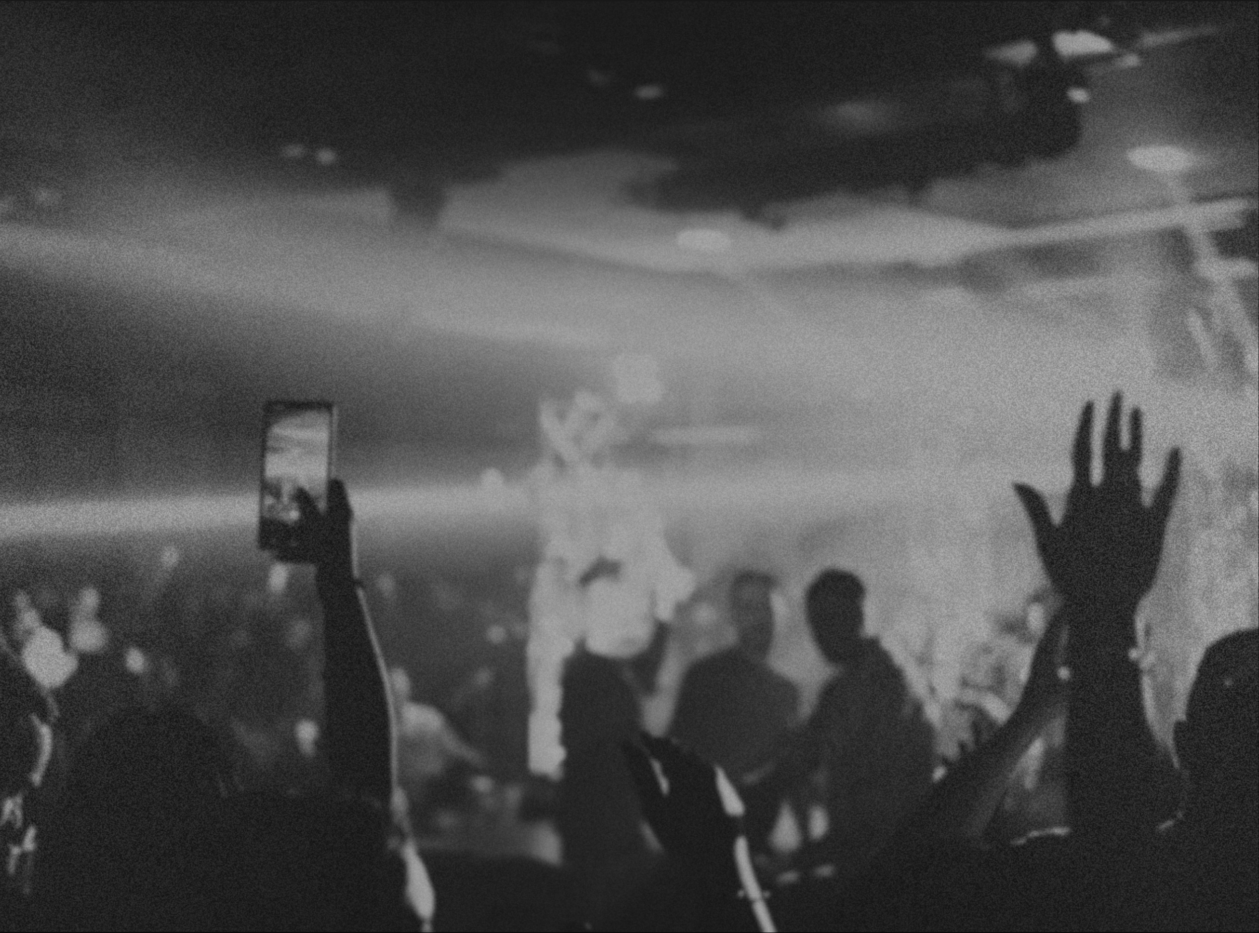 A black and white photo of a crowd at a concert or event, with one person holding a phone up to take a picture or video. The crowd appears to be waving or raising their hands, and the background shows a cloudy sky.