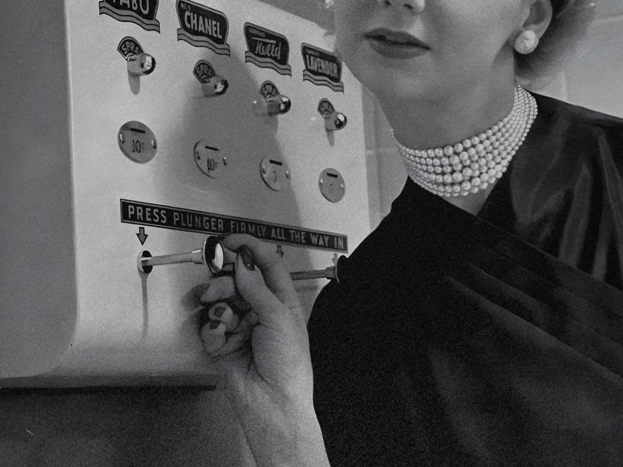 A woman wearing a pearl necklace and earrings is pressing a plunger button on perfume vending machine. The machine has various symbols and a label that reads 'Press plunger firmly all the way in'. Image rétro d'un distributeur automatique de parfum.