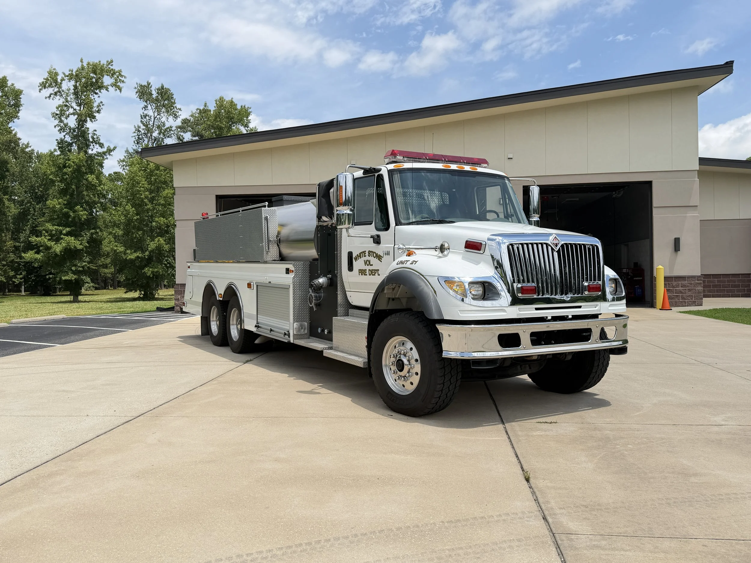 A white fire truck is parked in front of a fire station garage on a sunny day with blue sky and trees in the background.