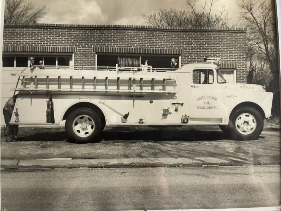 A black and white photo of a vintage fire truck parked in front of a brick building. The truck has a ladder, hoses, and firefighting equipment on top and in the side compartments. The side of the truck reads 'Gothic Fire Vol. Vire Dept.' A person is standing near the back of the truck.
