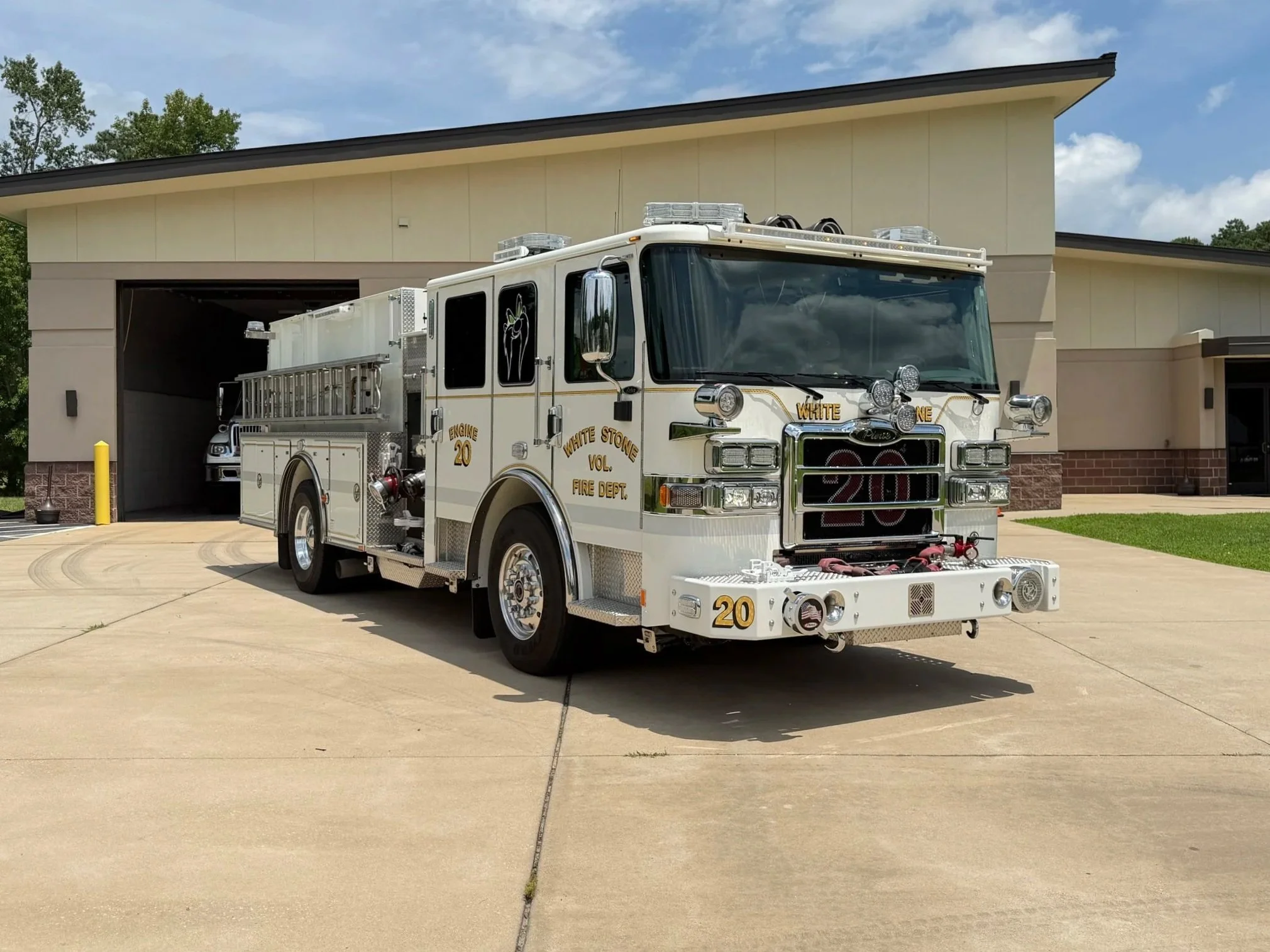 A white fire truck from the White Stone Volunteer Fire Department parked in front of a building with a garage door, under a partly cloudy sky.