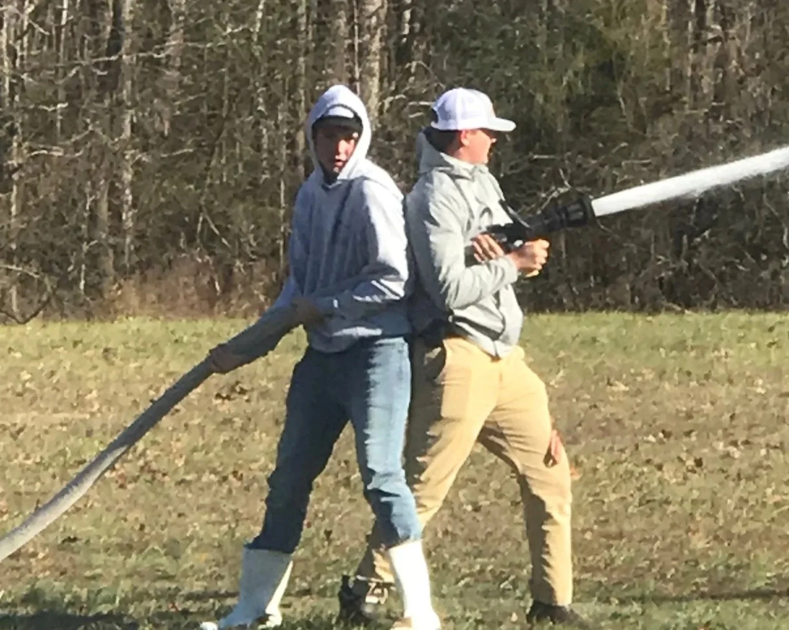 Two boys are outdoors, one holding a hose spraying water, and the other standing behind him.