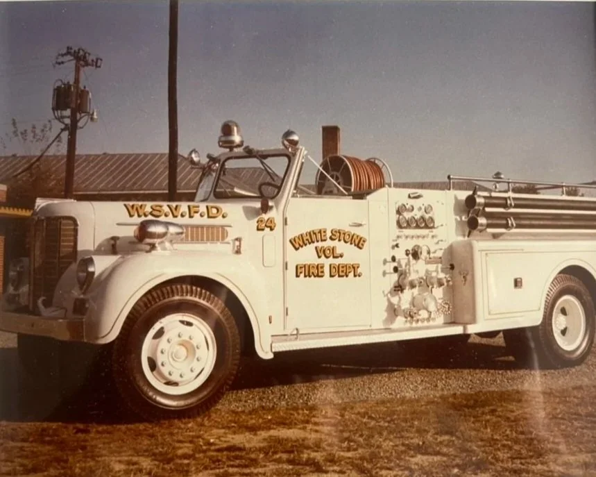 An antique white fire truck with the words 'White Stone Vol. Fire Dept.' on the side and 'W.S.V.F.D.' on the front, parked outdoors on dirt ground with a partly cloudy sky.
