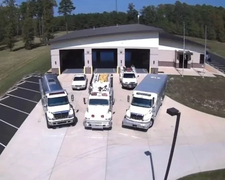 Three large trucks parked in front of a modern building, with a smaller white vehicle and a utility truck nearby.