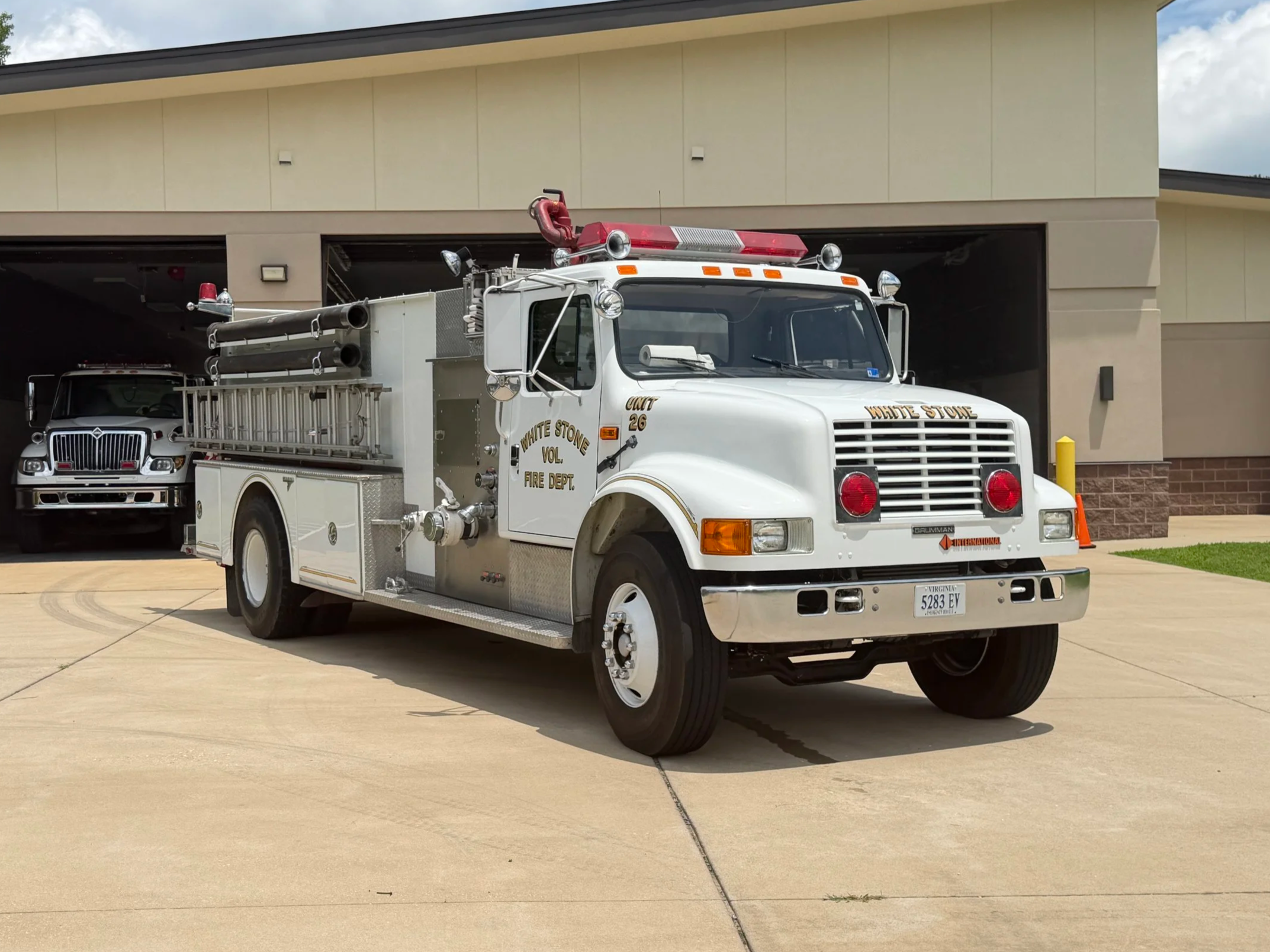 A fire truck with white and red coloring parked outside a building with a tan and beige facade, featuring large garage doors, in a paved area.