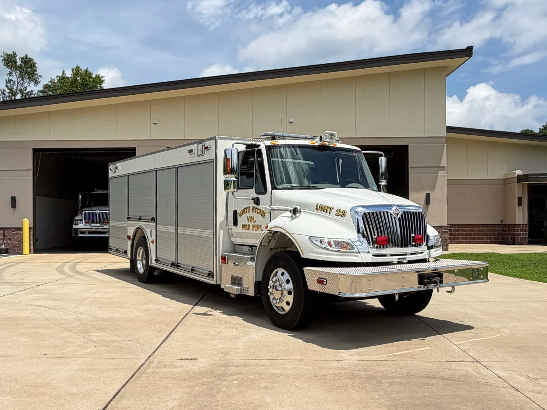 A white fire department rescue truck parked in front of the fire station, with a fire truck visible inside the garage.