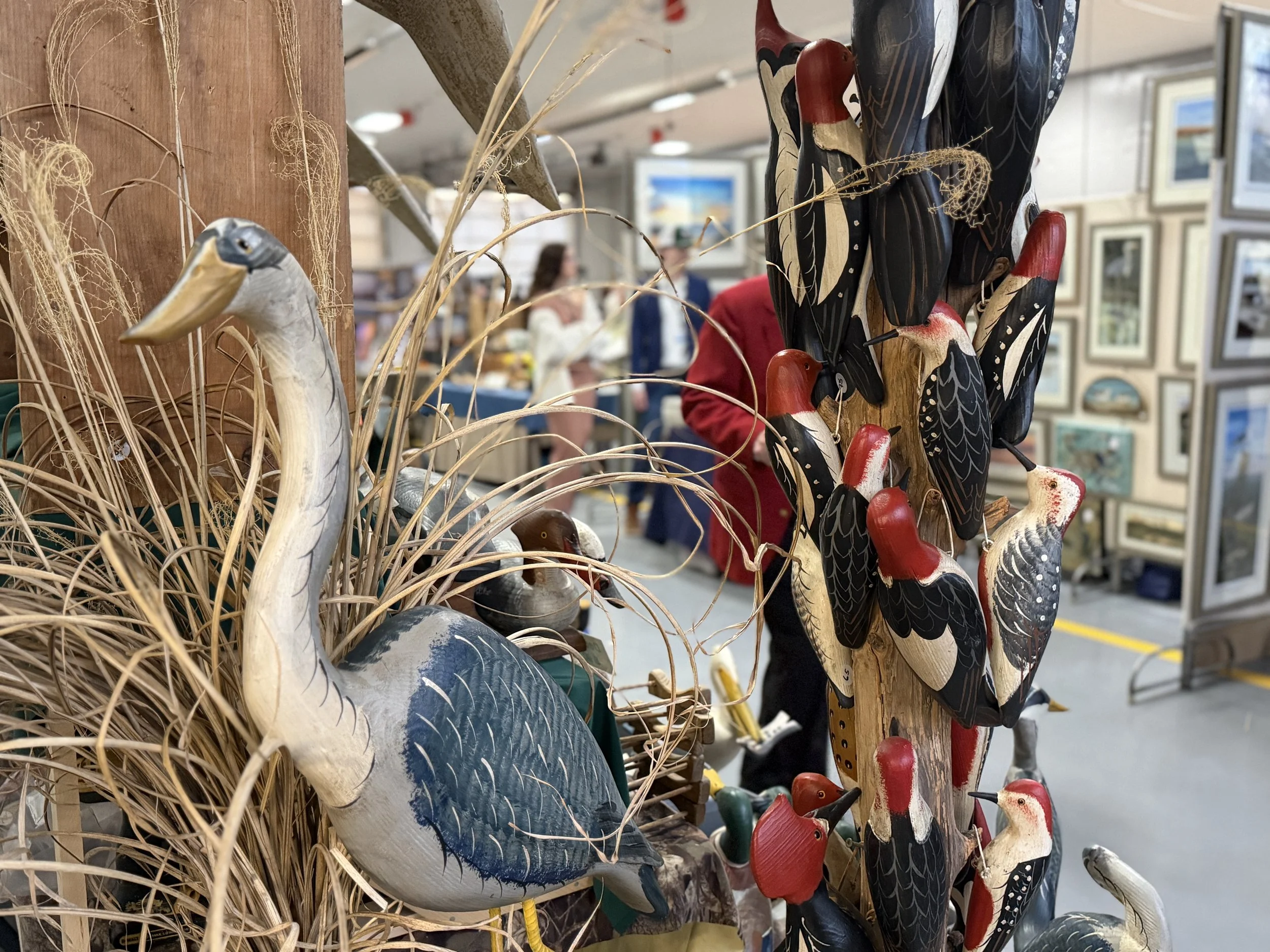 Decorative wood carvings of herons and ducks, with dried grass and twigs in the foreground, displayed at an indoor marketplace.