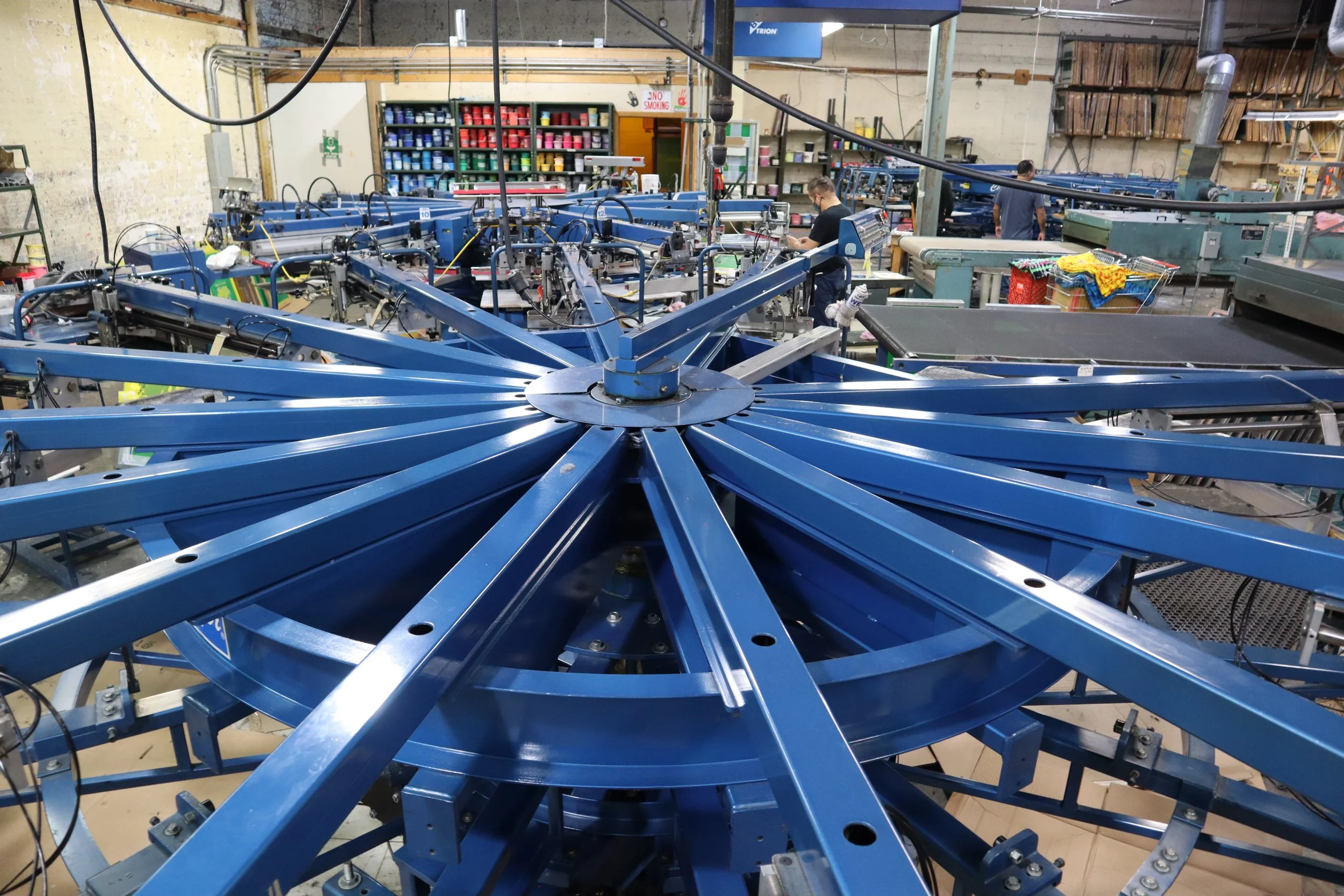 Interior of a manufacturing or factory workshop with multiple blue industrial machines, workers, and shelves filled with colorful materials.