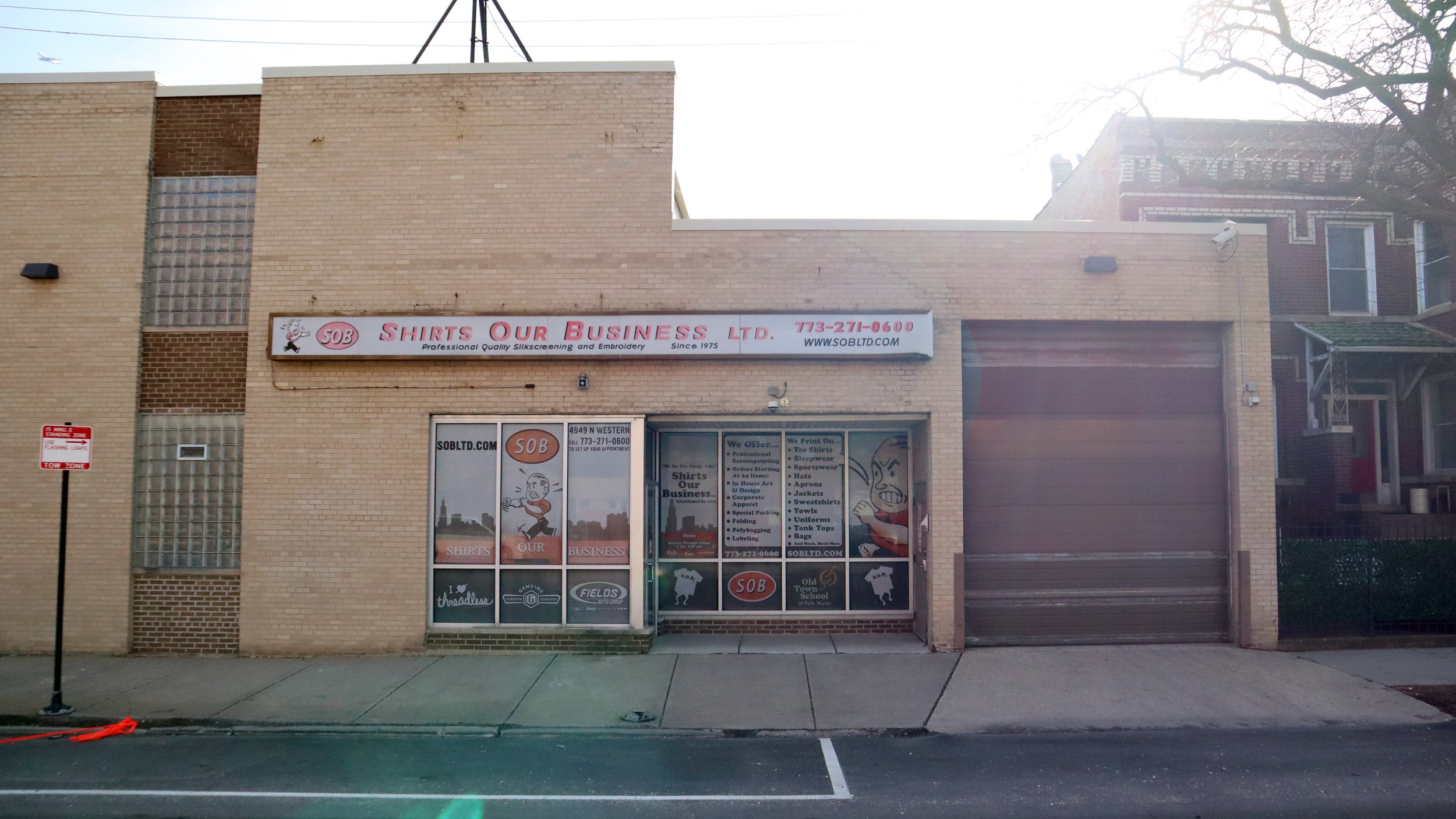 Storefront for 'Shirts Our Business Ltd.', a shirt printing and embroidery business, with signage displaying contact information and services offered, window graphics, and a large garage door.