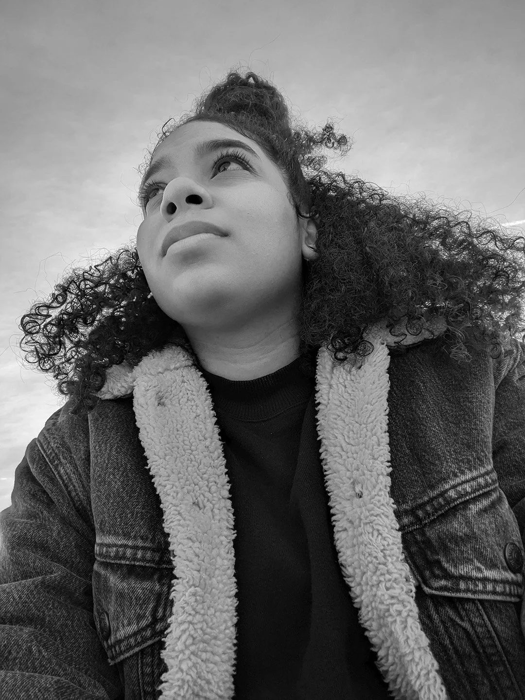 Black-and-white photo of Rosa outdoors, looking upward with a thoughtful expression, wearing a jacket with a sherpa lining.