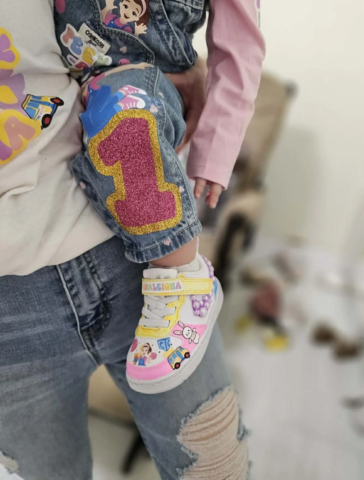 Close-up of a child being carried, wearing colorful sneakers with decorated illustrations and the name "Malebria" on a strap, ripped jeans, and a pink long-sleeve shirt. The child’s denim pants have a large, glittery pink and gold number one patch.