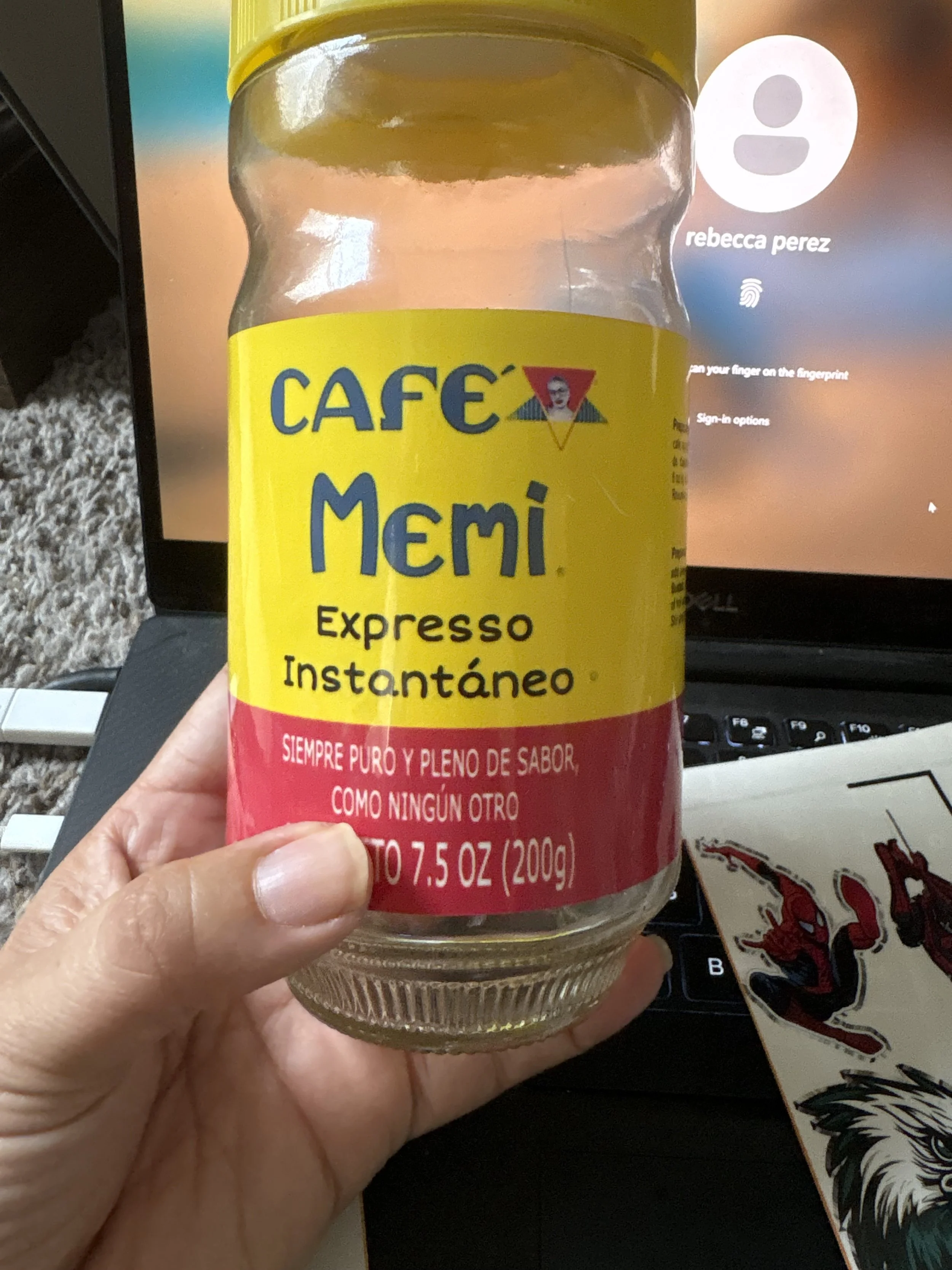 A glass jar of Café Memi instant espresso, with yellow and red label, held in front of a laptop. The label states it contains 7.5 ounces (200 grams) and is described as pure and full of flavor.