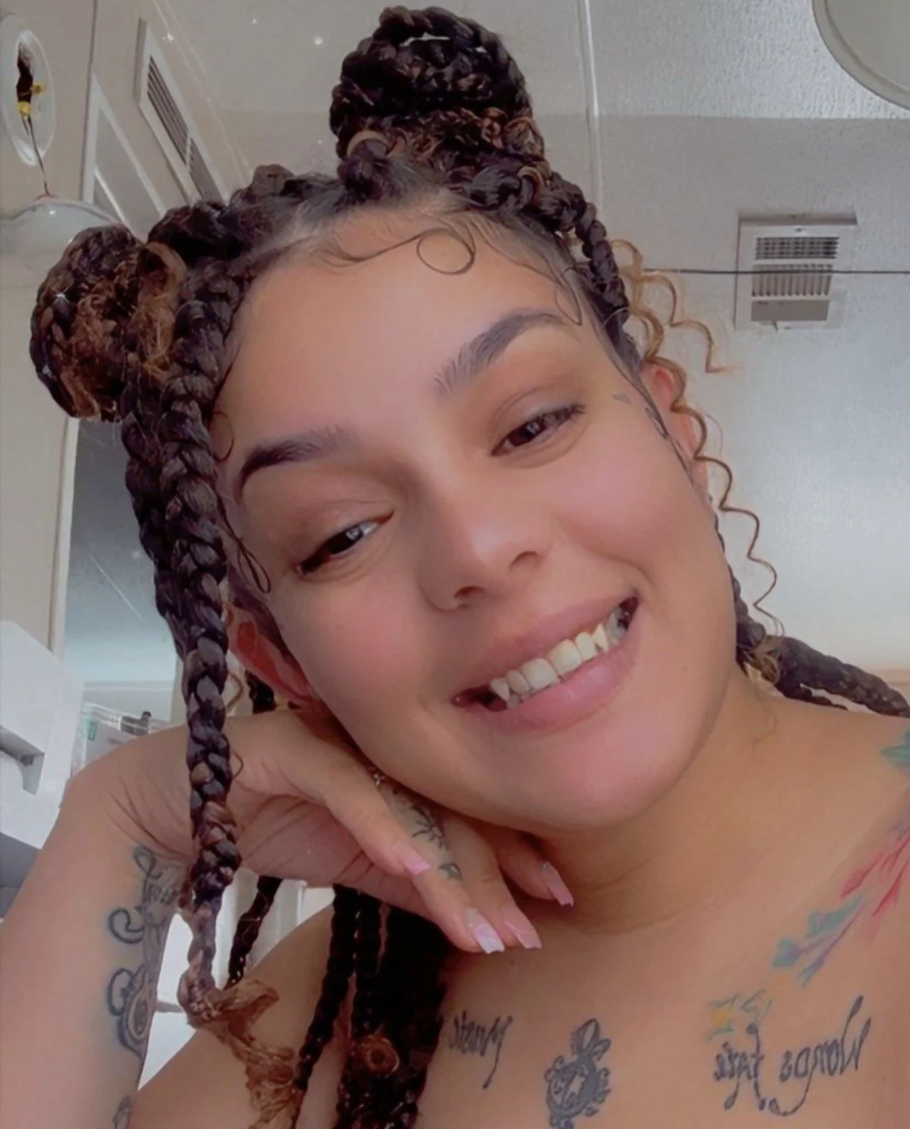 Close-up of a woman smiling with tattoos visible on her shoulder and chest, and her hair styled in braids and buns.