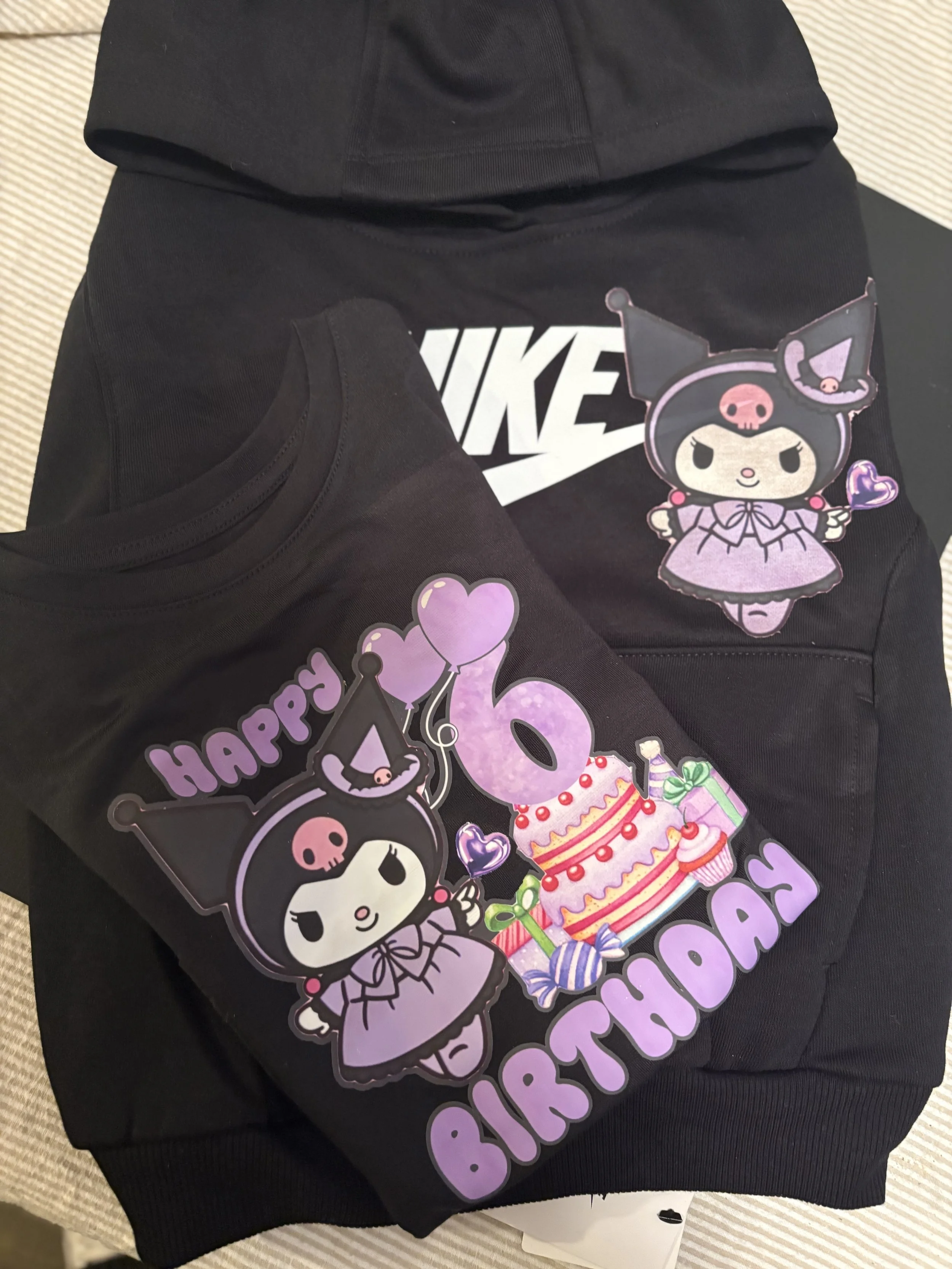 Black Nike hoodie on top of black sweatshirt with Happy 16th Birthday decorations featuring a cute girl with cat ears, a birthday cake, and heart-shaped balloons, all in purple and pink colors.