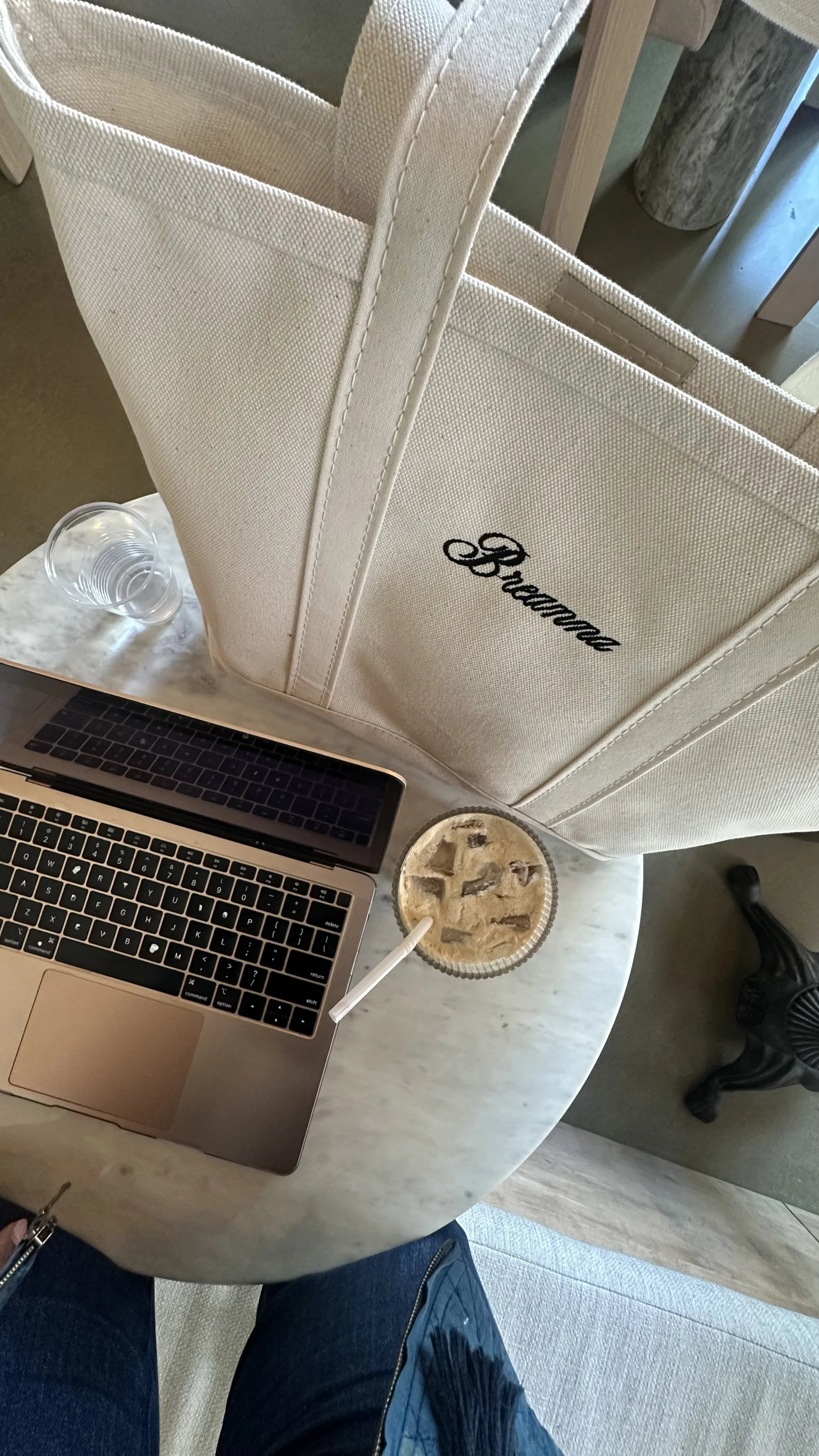 A marble table with a laptop, an iced coffee with a straw, an empty glass, and a large beige tote bag with the word 'Browse' embroidered on it, taken from a top-down perspective.