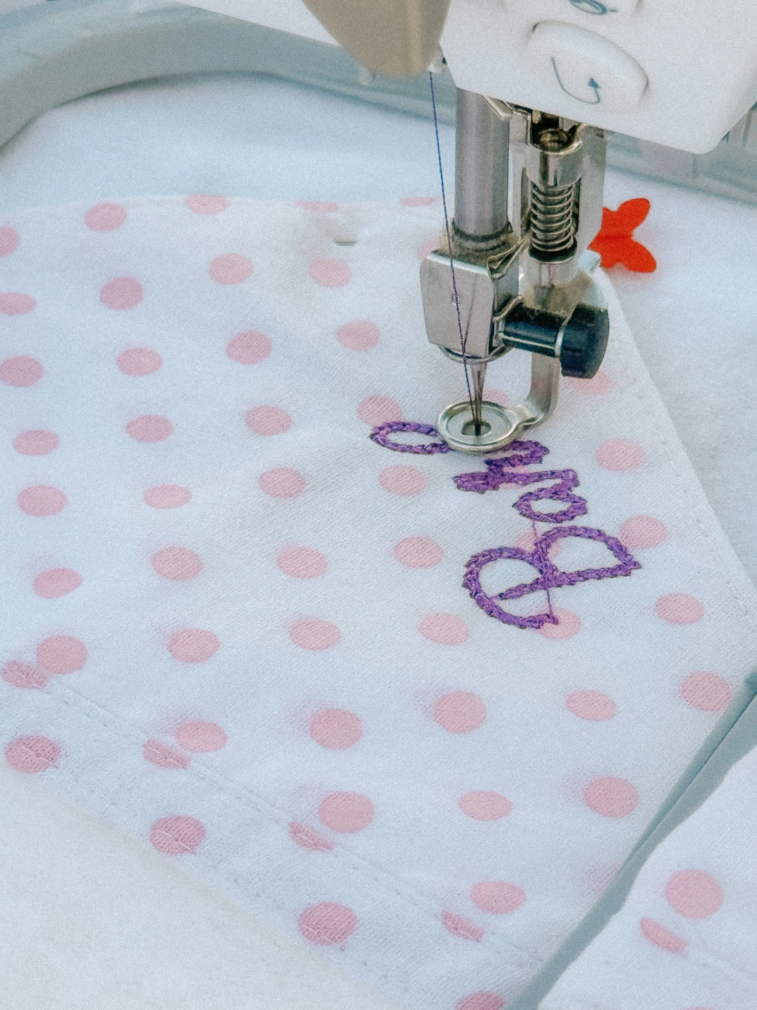 Close-up of a sewing machine embroidering the word 'baby' in purple thread on white fabric with pink polka dots.