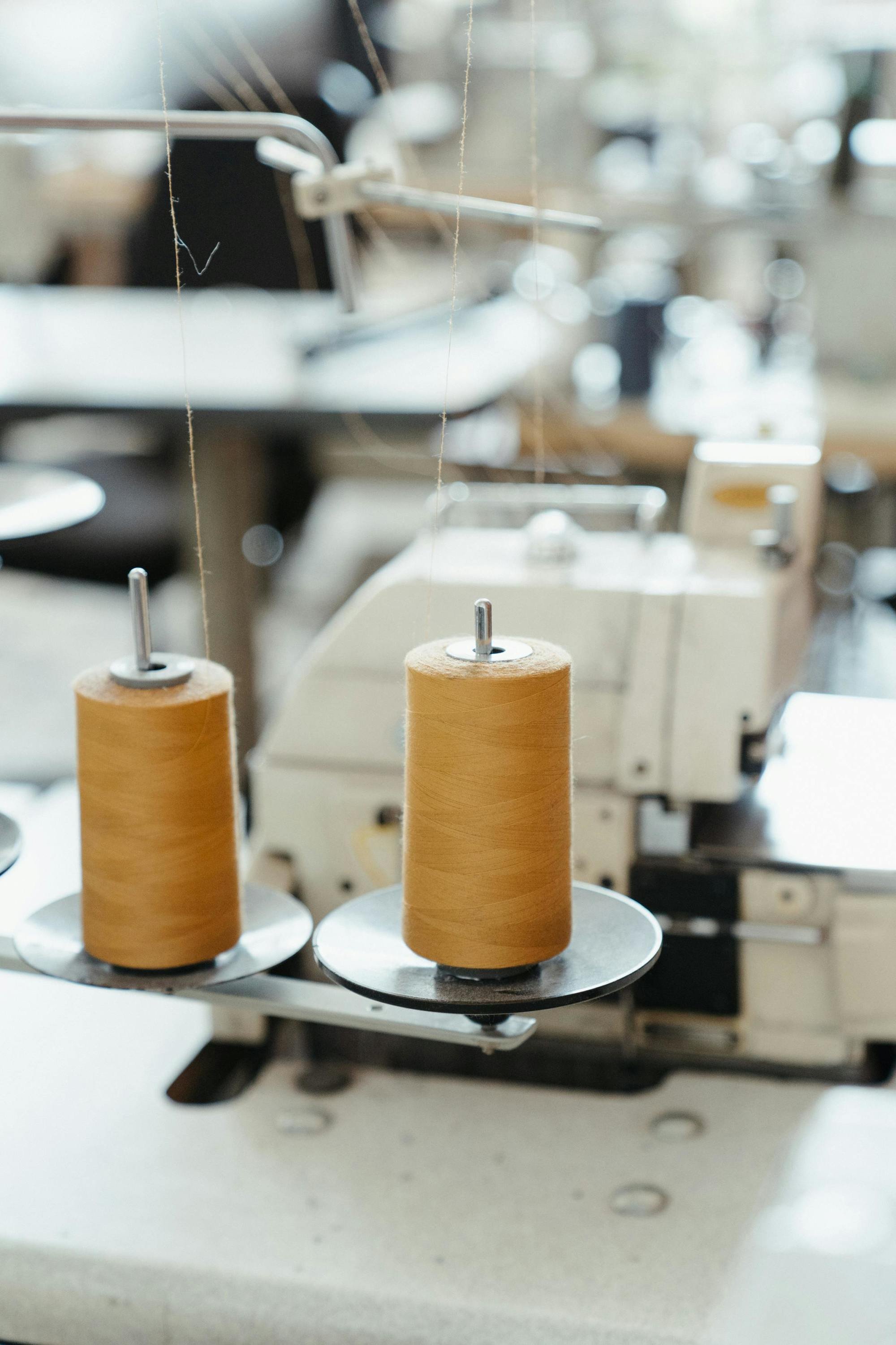 Close-up of a textile sewing machine with spools of beige thread in a factory or workshop setting.