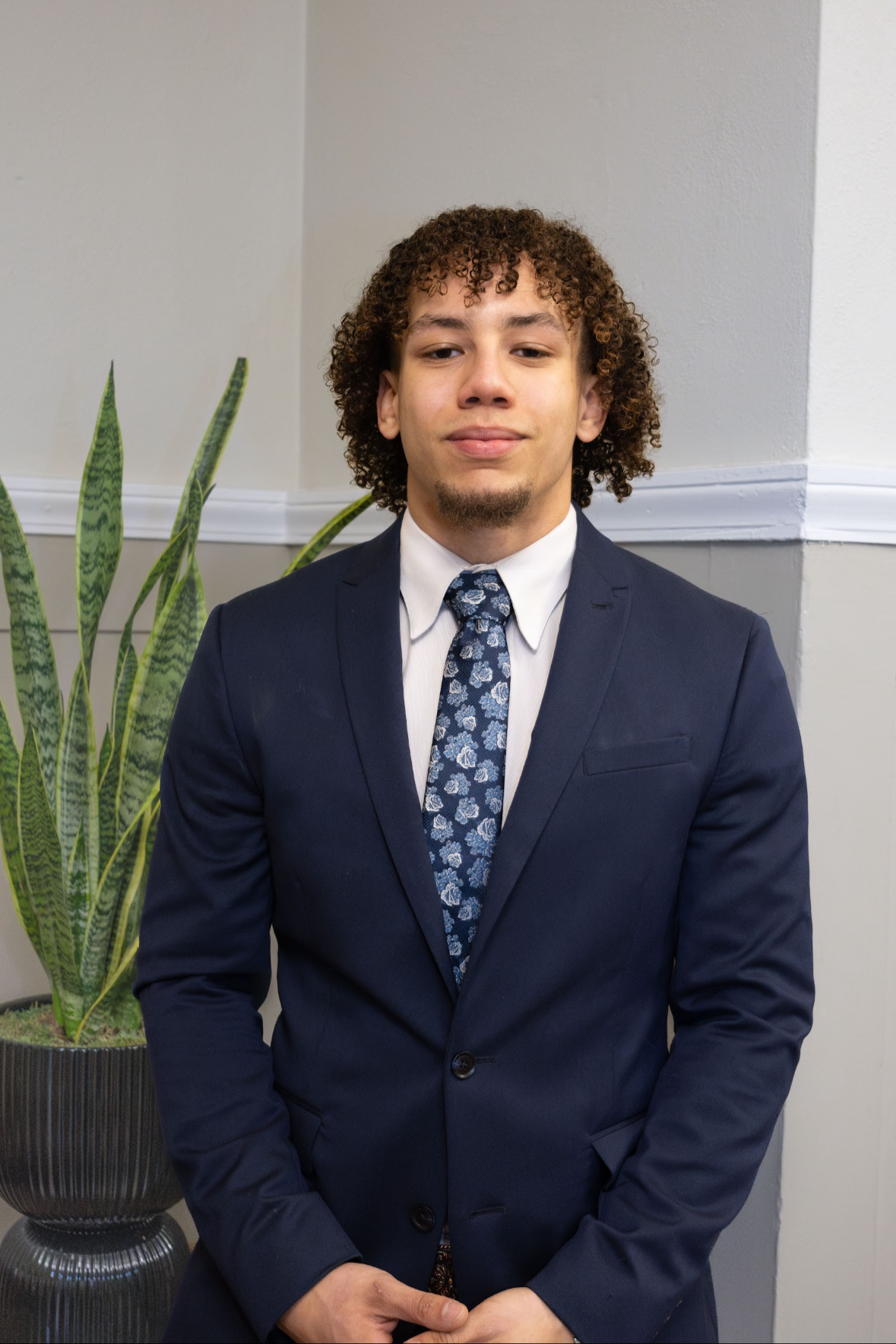Young man in a navy blue suit, white shirt, and floral tie posing indoors near a potted snake plant.