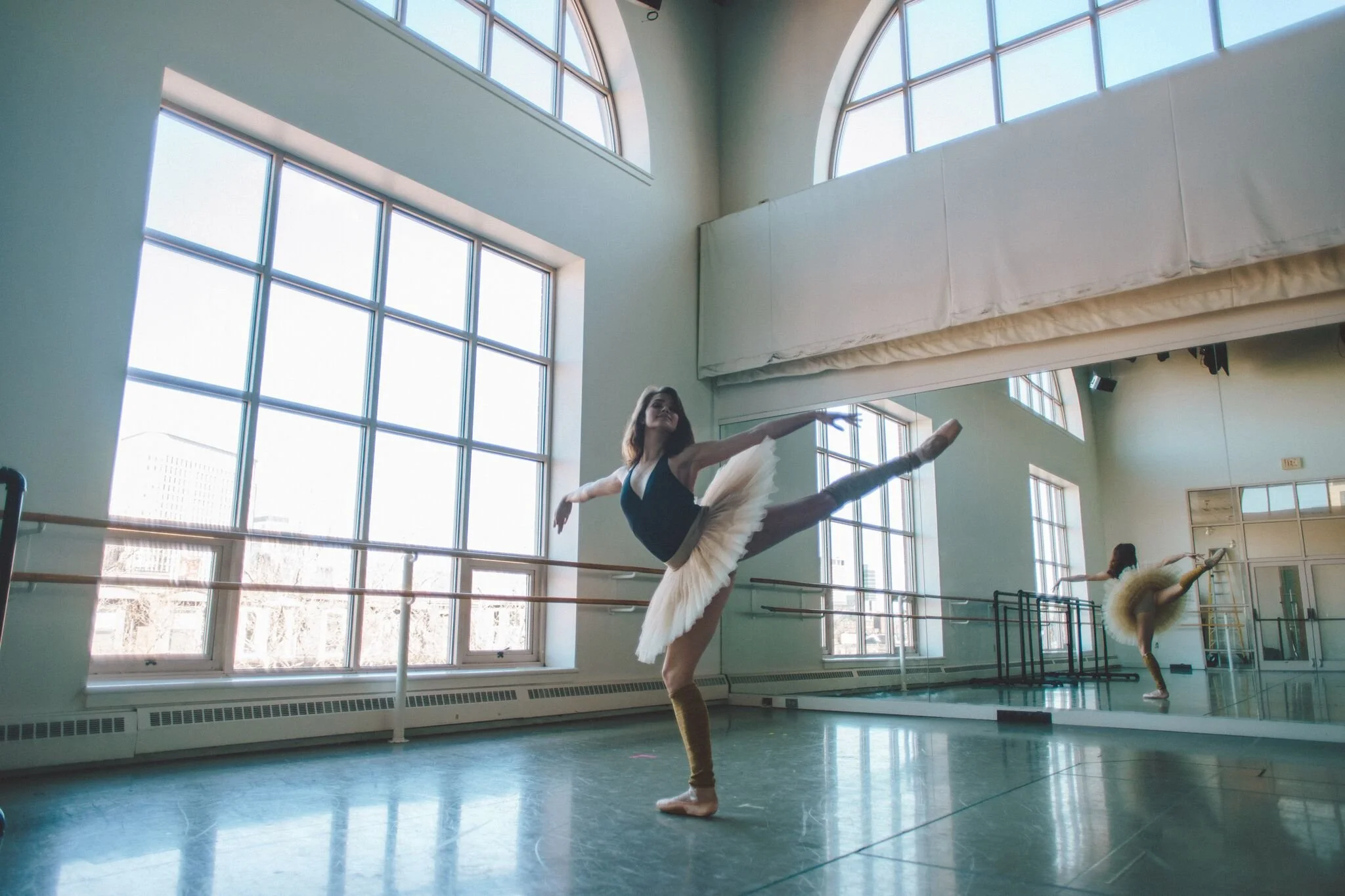 A ballerina in a studio performing an arabesque pose while stretching her arm and leg, dressed in a black leotard and a white tutu.