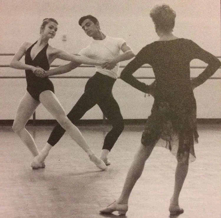 Two ballet dancers practicing for an instructor in a dance studio.  The dancers are partnered, holding hands, while the instructor observes.