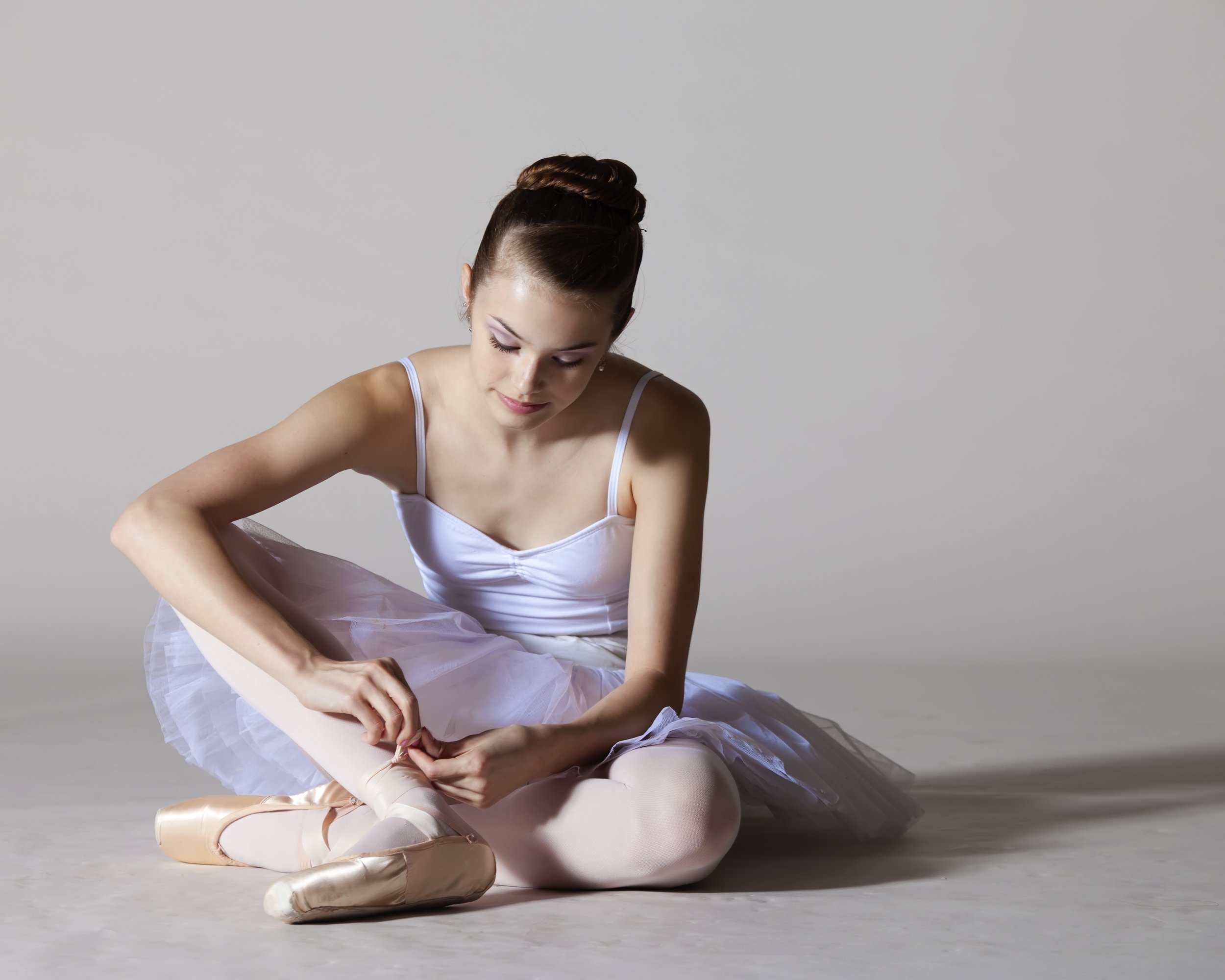Ballet dancer in a white tutu and ballet slippers sitting on the floor, adjusting her pointe shoe against a plain gray background.