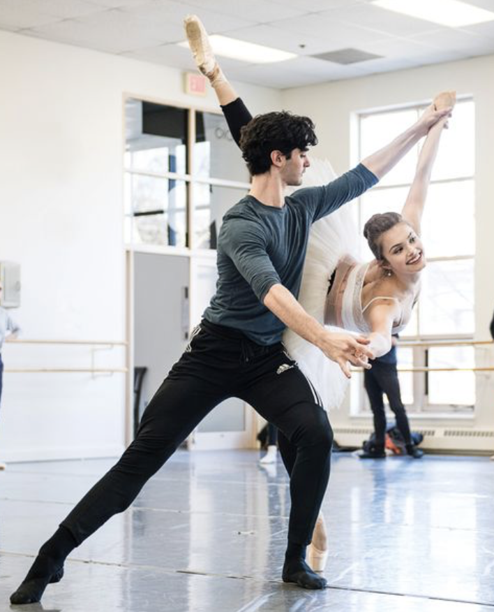 A ballet dancer practicing with an instructor in a dance studio, with the dancer executing a split pose and the instructor assisting.