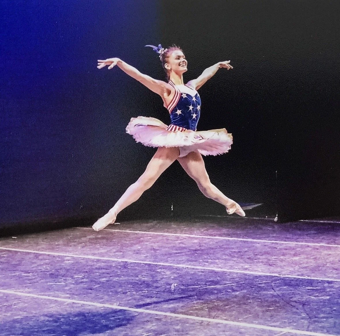 Ballet dancer performing on stage in a patriotic-themed costume with star and stripe patterns, jumping with arms and legs outstretched in a star shape.