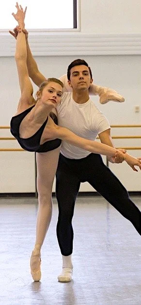A ballerina and a male ballet dancer during partnering rehearsal in a studio with large windows and ballet barres.