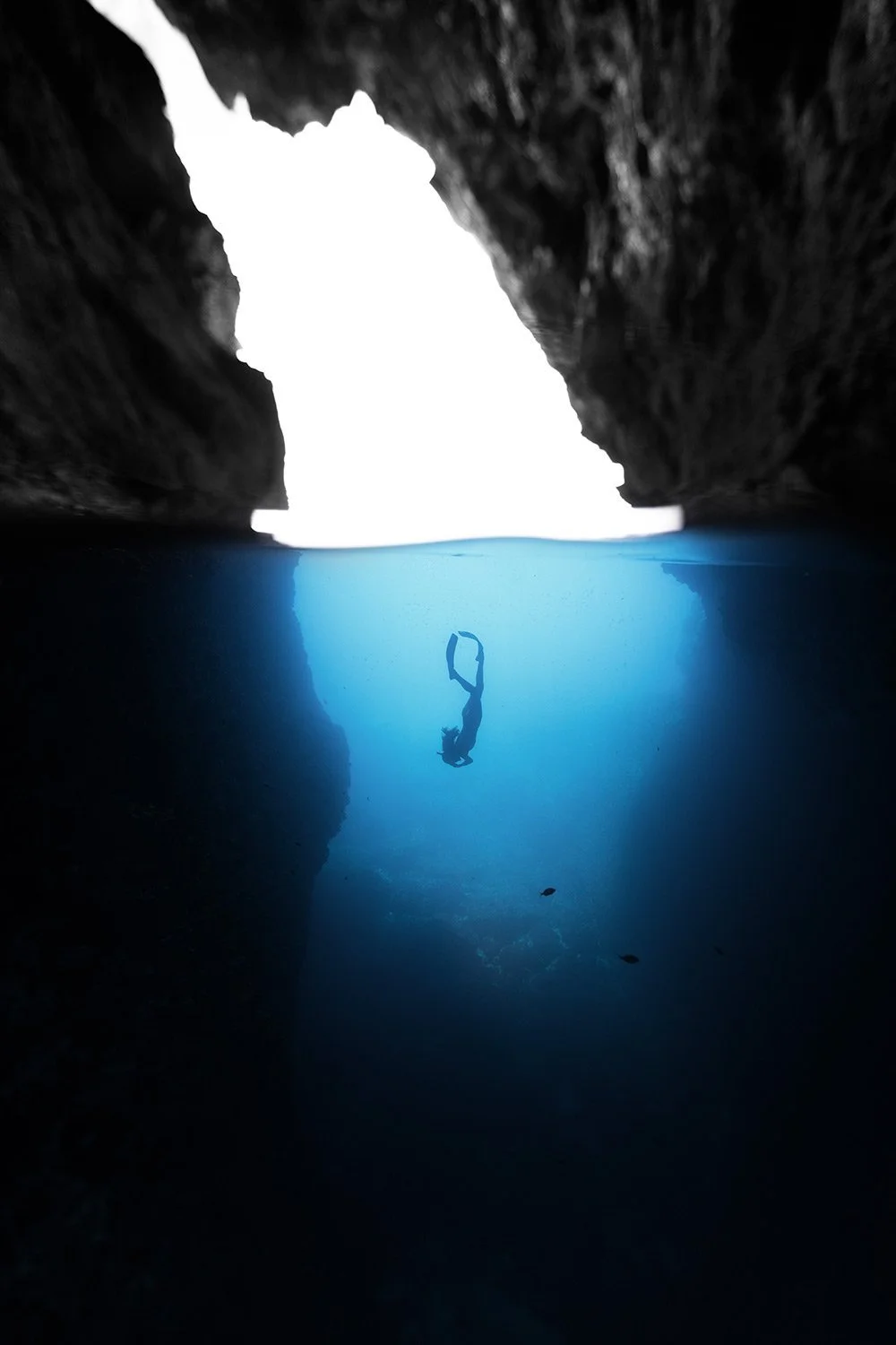 A person snorkeling in a narrow sea cave with an opening to the outside, viewed from beneath the water.