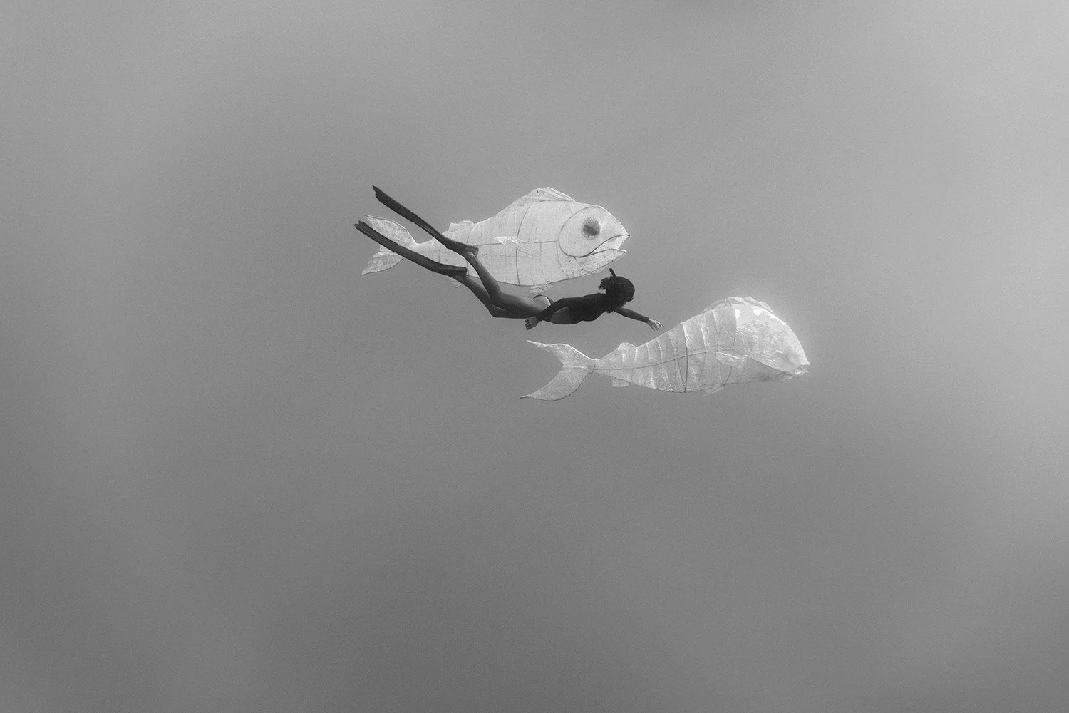 A person appears to be flying through the sky with wings made of paper, alongside two fish-shaped paper lanterns.