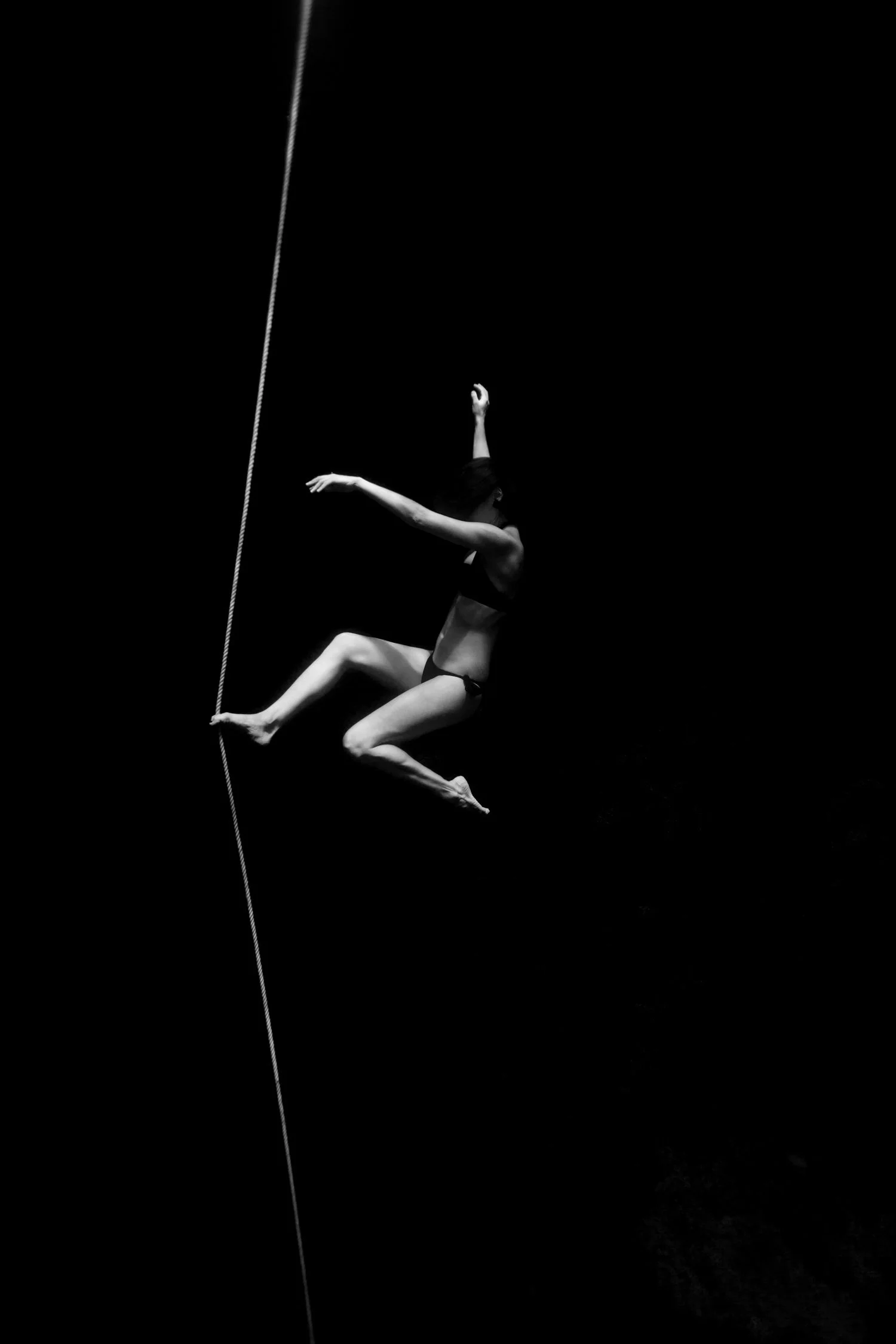 A black and white photo of a dancer mid-performance, suspended in the air, holding a thin rope, with a dark background.