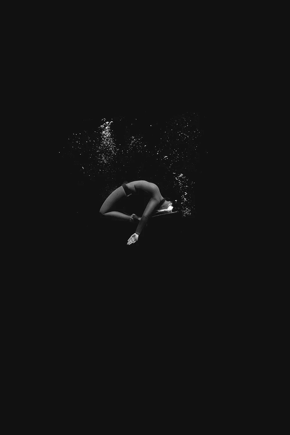 A person in a black wetsuit is underwater, curled in a fetal position, with bubbles rising around them.