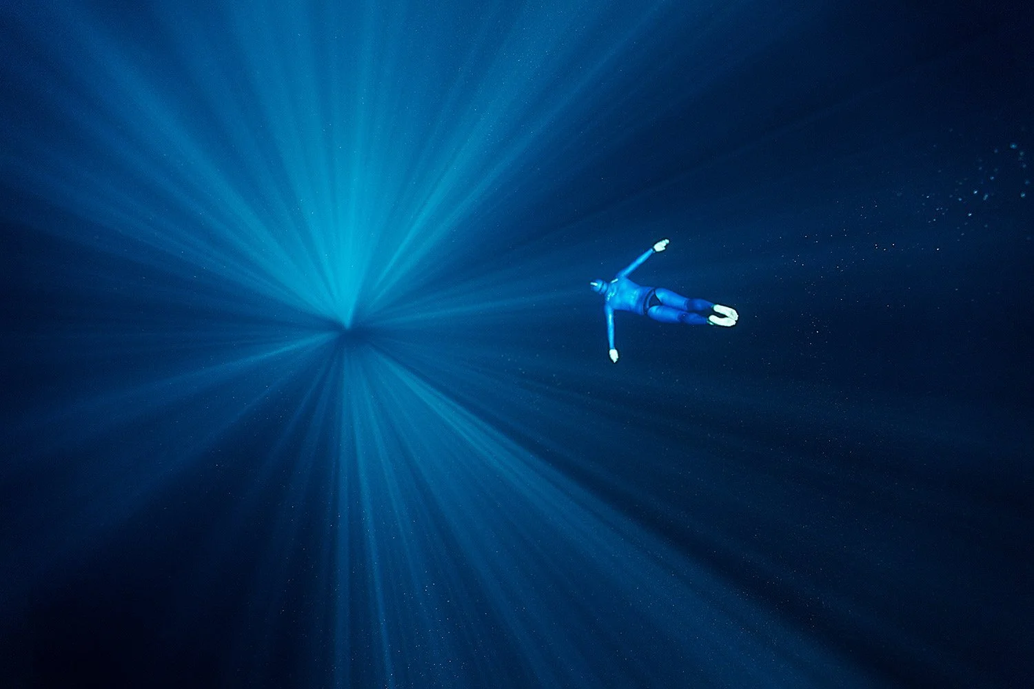 A person in a wetsuit swimming underwater towards a bright light, surrounded by deep blue water and small floating particles.