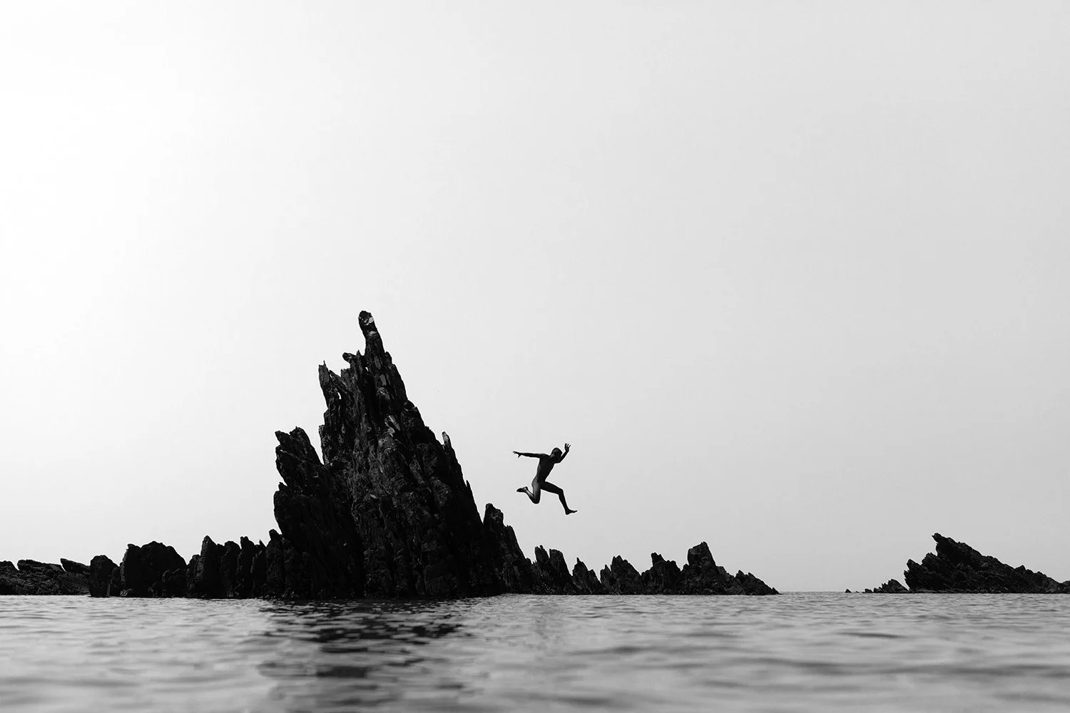 A person jumping off a jagged rock formation into the water, with a minimalist black and white landscape and a cloudy sky.