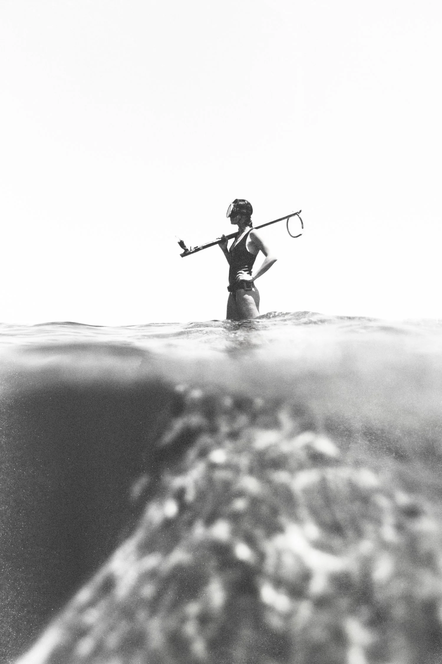 A person standing in the water, wearing a swimsuit and a helmet, holding a spear with a mask on.