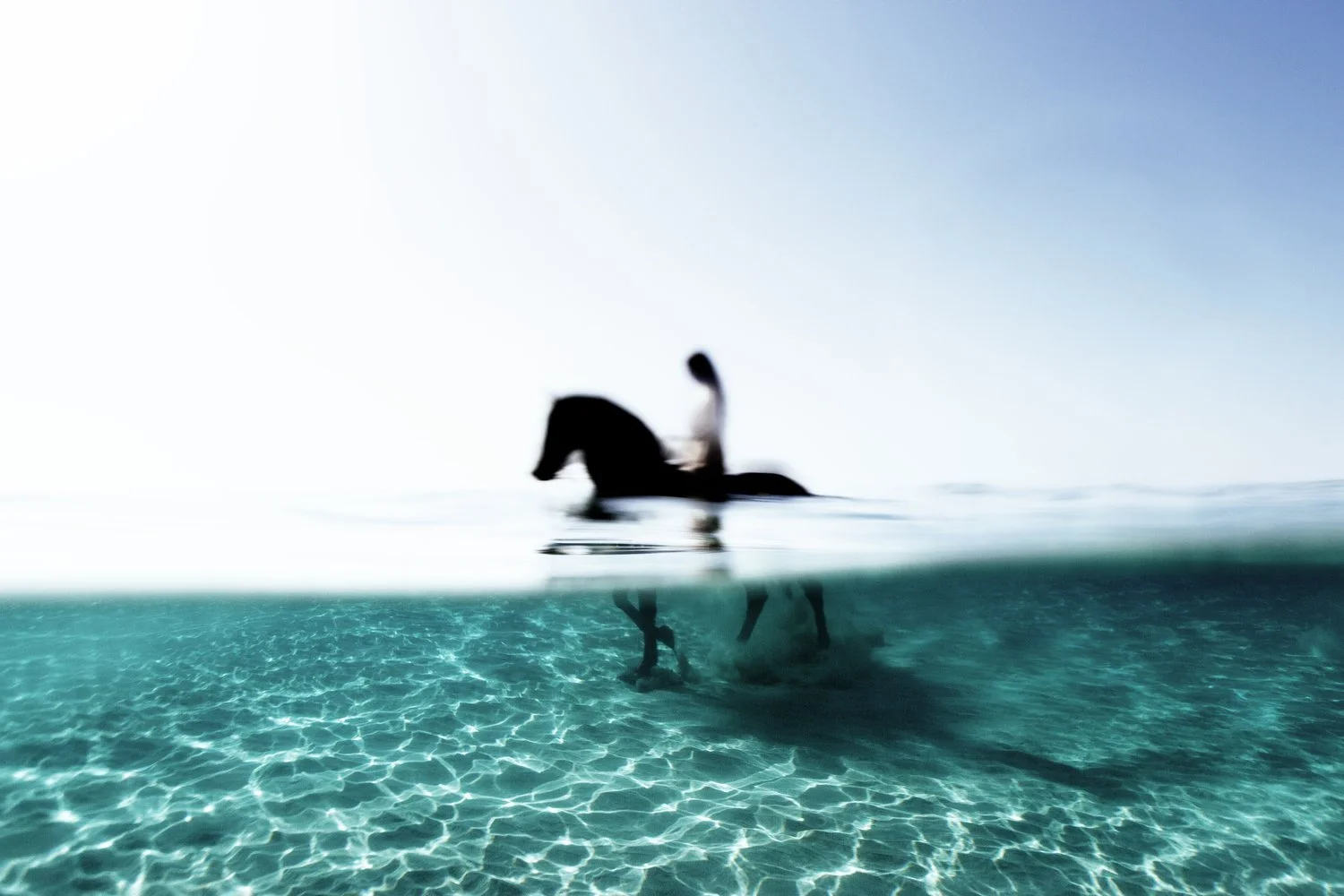 A person riding a horse in the ocean, with the underwater part of the horse and rider visible beneath the water's surface. The water is clear and turquoise, with the sky above being bright and clear.