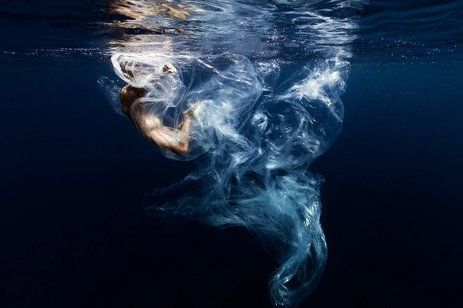A person underwater with their body curled, surrounded by flowing, semi-transparent plastic-like material.